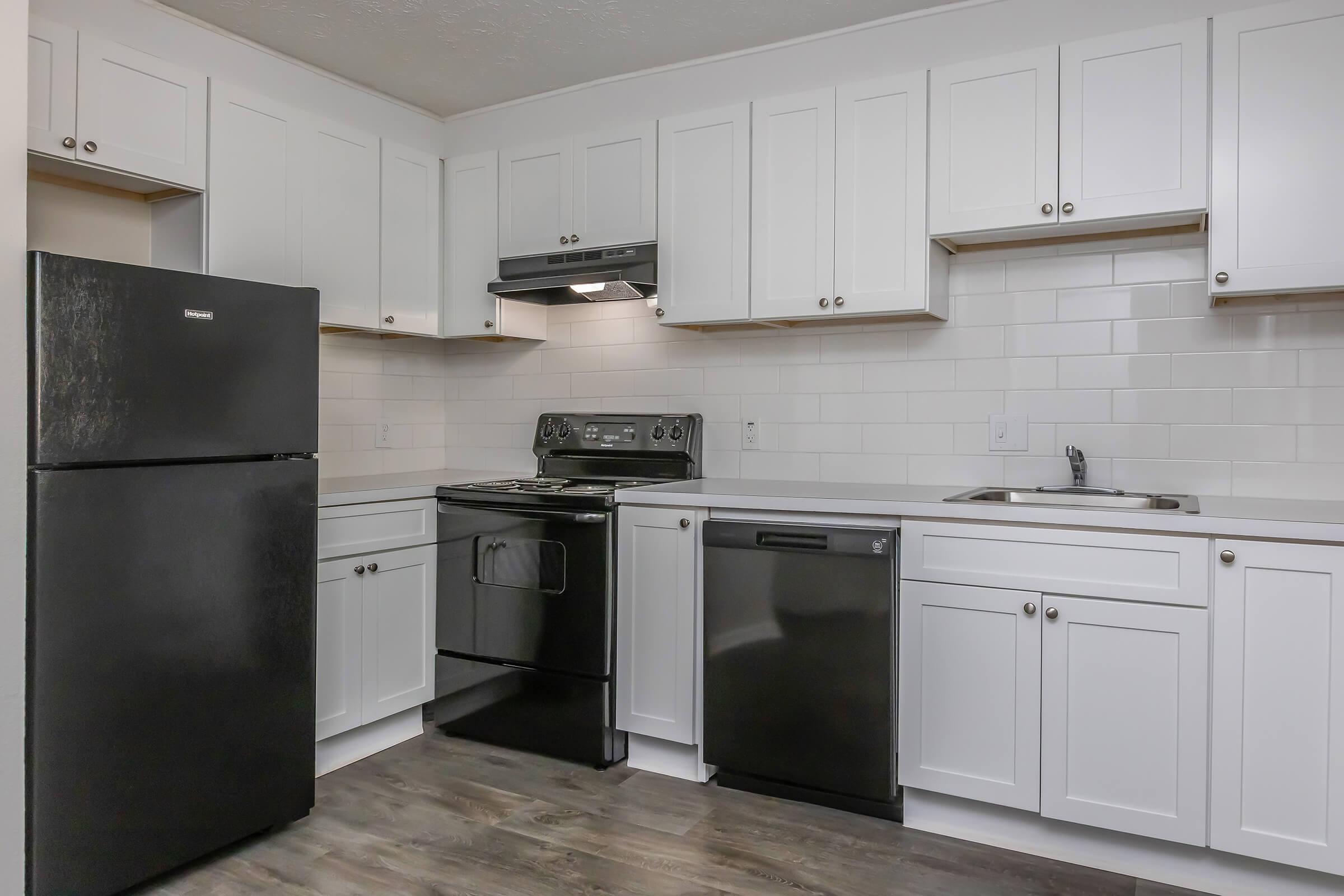 A modern kitchen featuring white cabinets, a black refrigerator, stove, and dishwasher. The countertops are light-colored, and there is a tile backsplash. The space is well-lit, showcasing a clean and organized layout, with a spacious feel and laminate flooring.