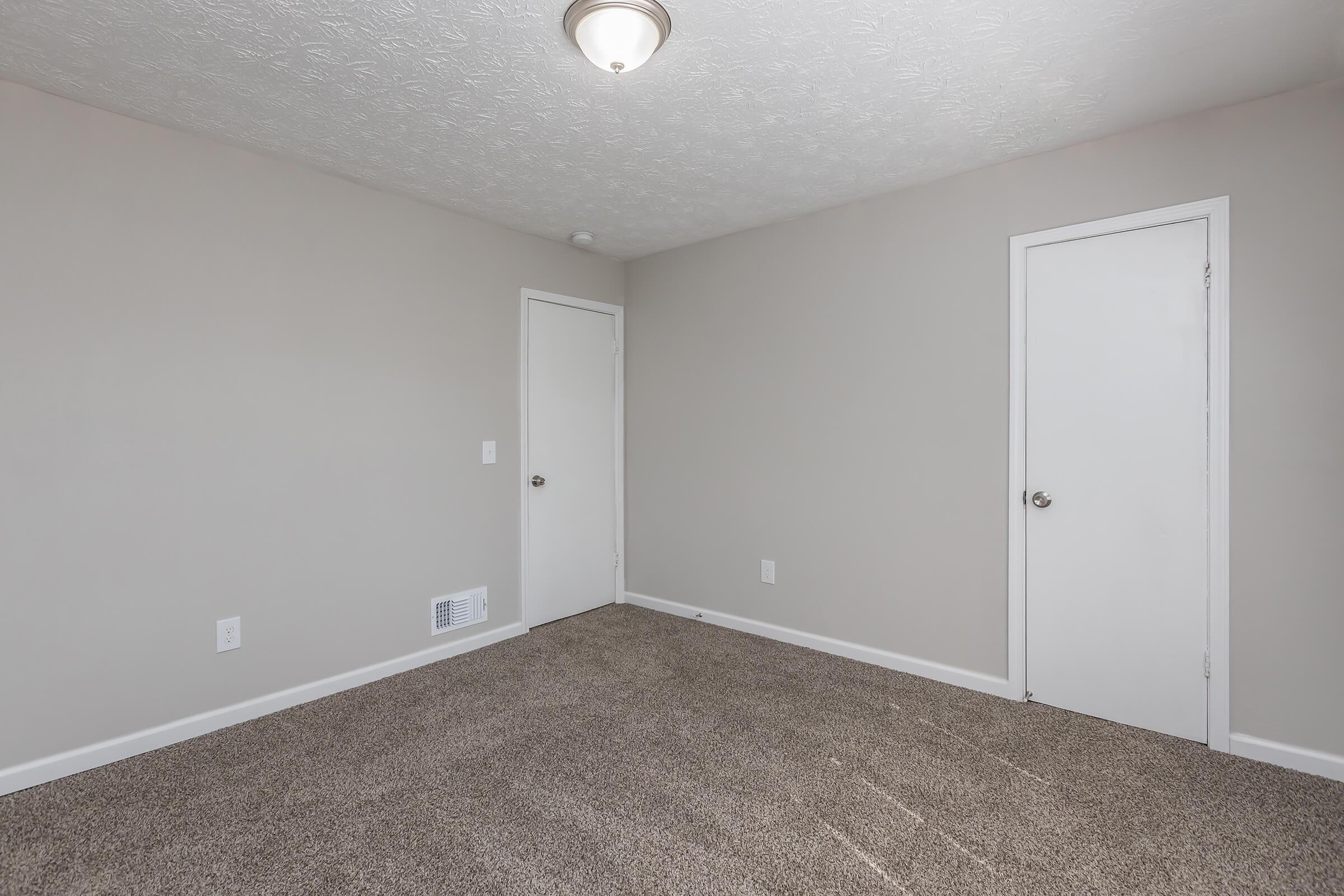 Empty room with light beige walls and a textured ceiling. The floor is covered with light brown carpet. There are two doors, one on the left and one on the right, and a ceiling light fixture. A small vent is visible on the wall near the floor. The overall ambiance is clean and minimalistic.