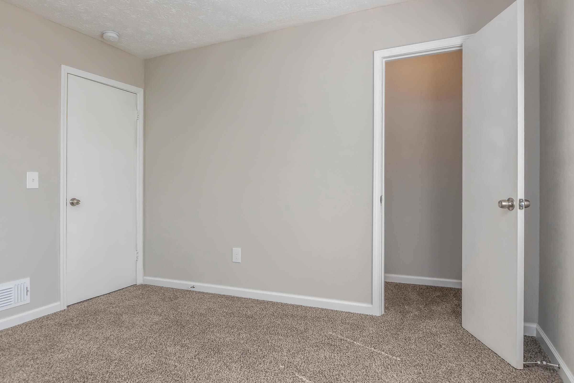 A simple room with light gray walls and beige carpet. One door leads to a closet, while another door is slightly open, revealing an empty space beyond. The room has no furniture and features a ceiling light fixture.