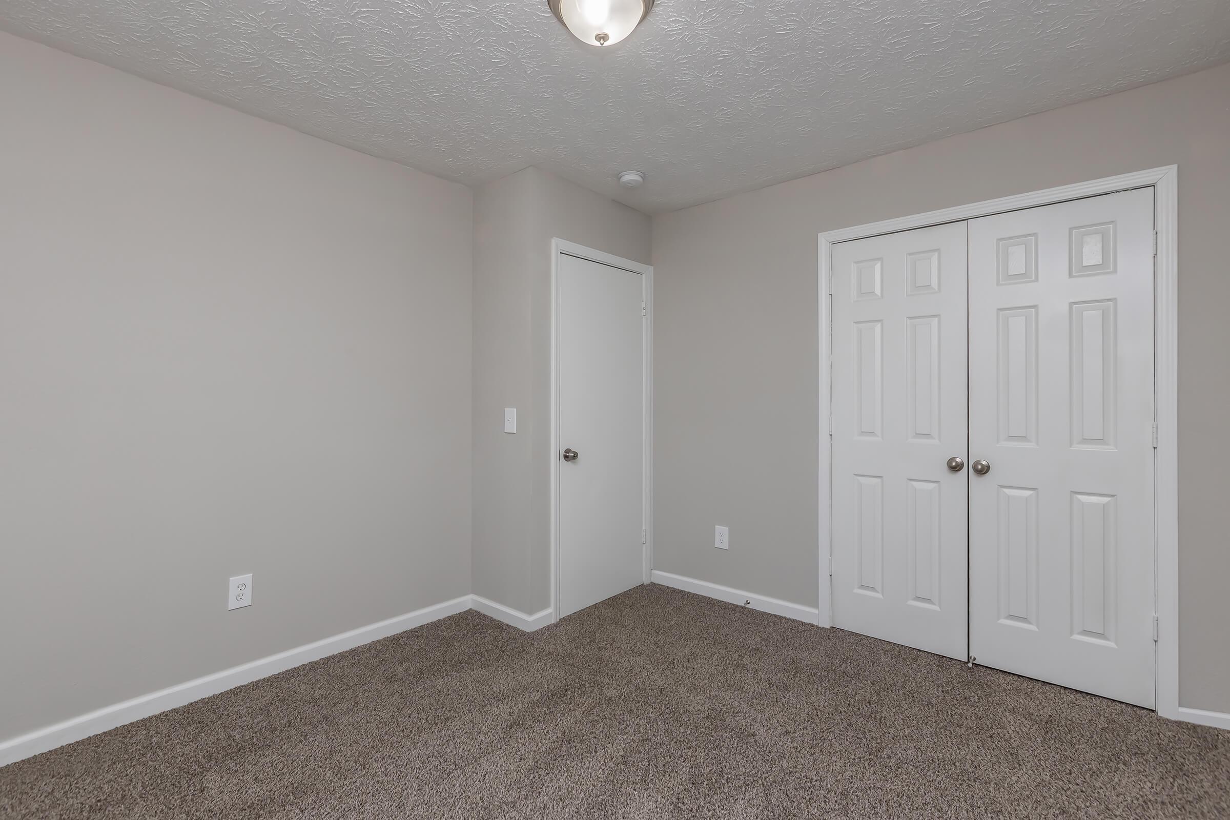 Interior of a light-colored bedroom with beige carpet. The walls are painted in a soft gray shade. There is a single door on the left and a double closet door on the right, both white. A ceiling light fixture is visible above, illuminating the space, which appears empty and well-maintained.
