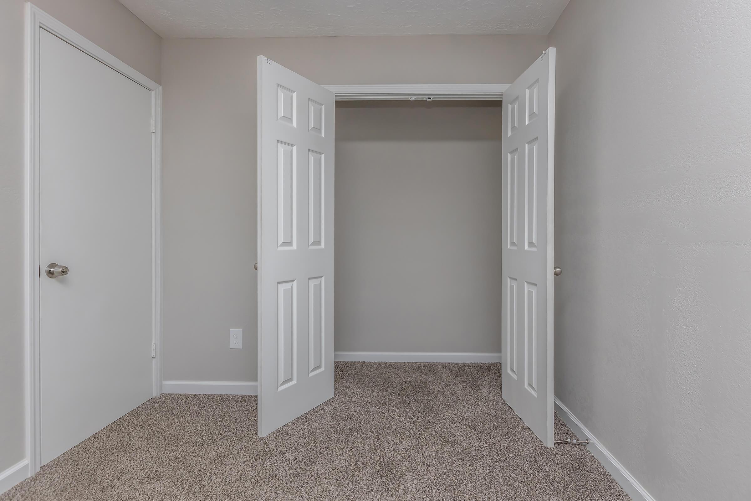 A view of an empty room with beige walls and carpeted flooring. Two white double doors are open, leading to a small closet on the left, while a plain white door, likely leading to another room, is visible on the right. The lighting is bright and the space appears clean and well-maintained.