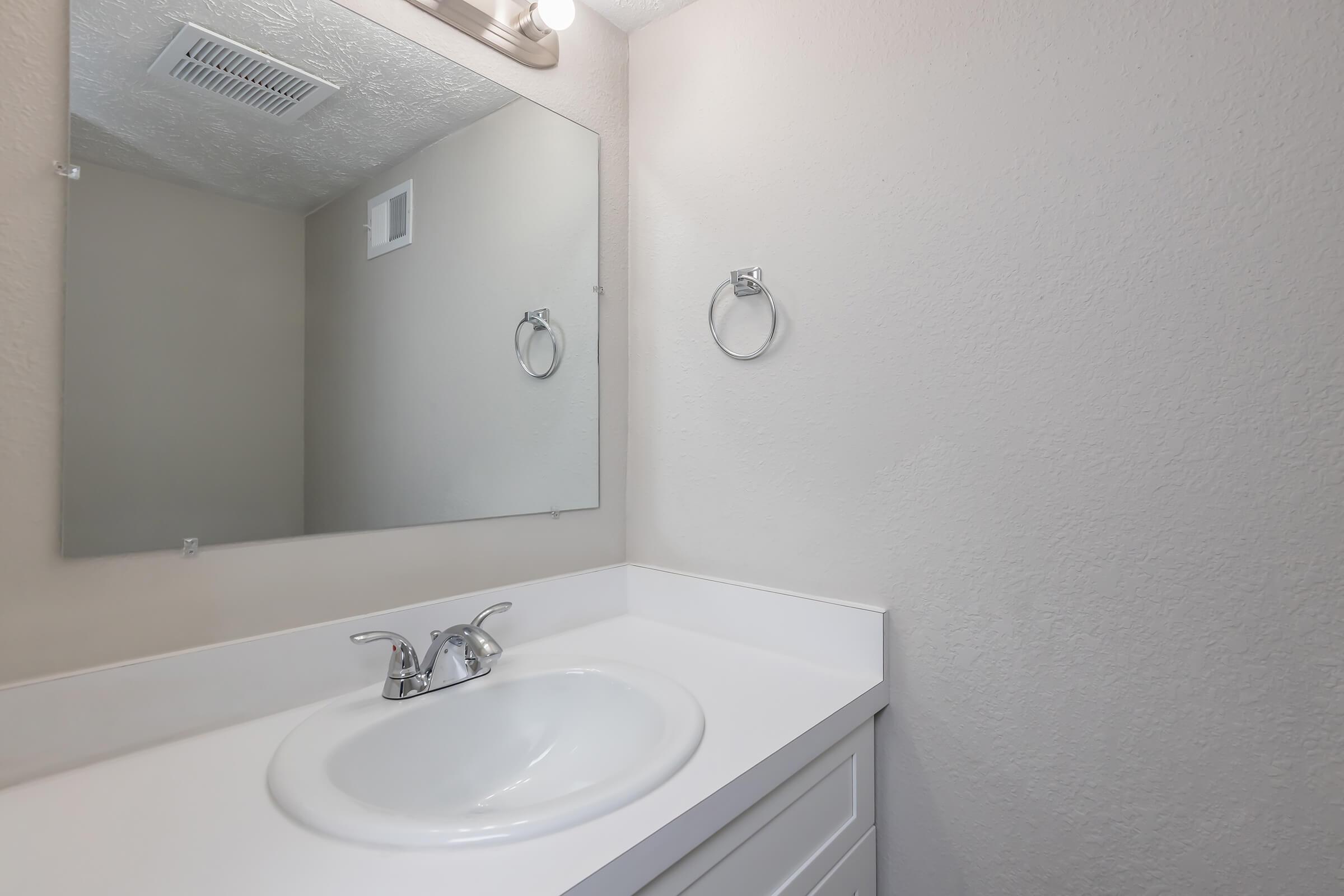 A clean, simple bathroom scene featuring a white sink with a chrome faucet, a round mirror above the sink, and a light fixture. The walls are a light neutral color, and a towel ring is mounted nearby. The overall ambiance is bright and minimalistic.