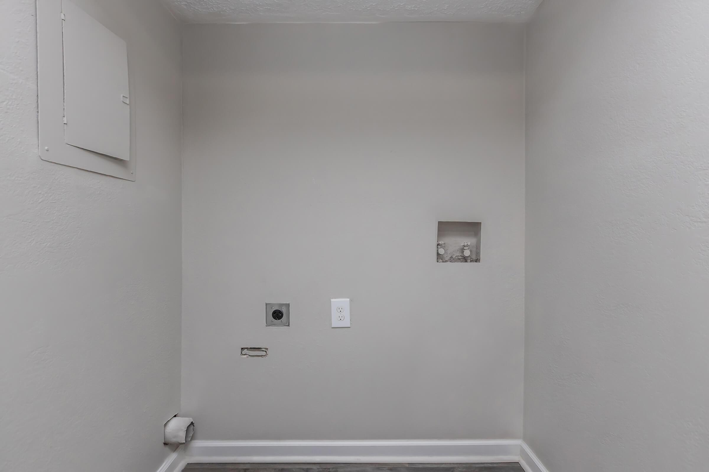 Empty room with light gray walls. A small square opening is visible on the right side, and another access panel is located on the left. The floor has a simple dark design, and there are no furnishings or decorations present in the space.