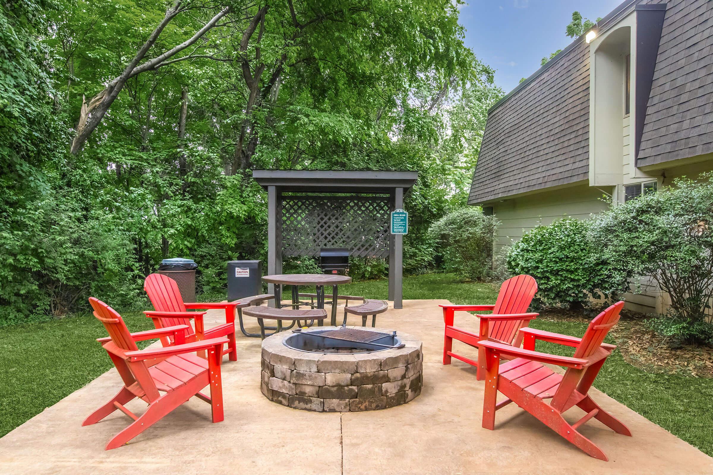 A cozy outdoor fire pit area featuring a circular stone fire pit surrounded by four red Adirondack chairs. There is a wooden shelter in the background, and lush greenery surrounds the space, creating a serene atmosphere for gatherings.