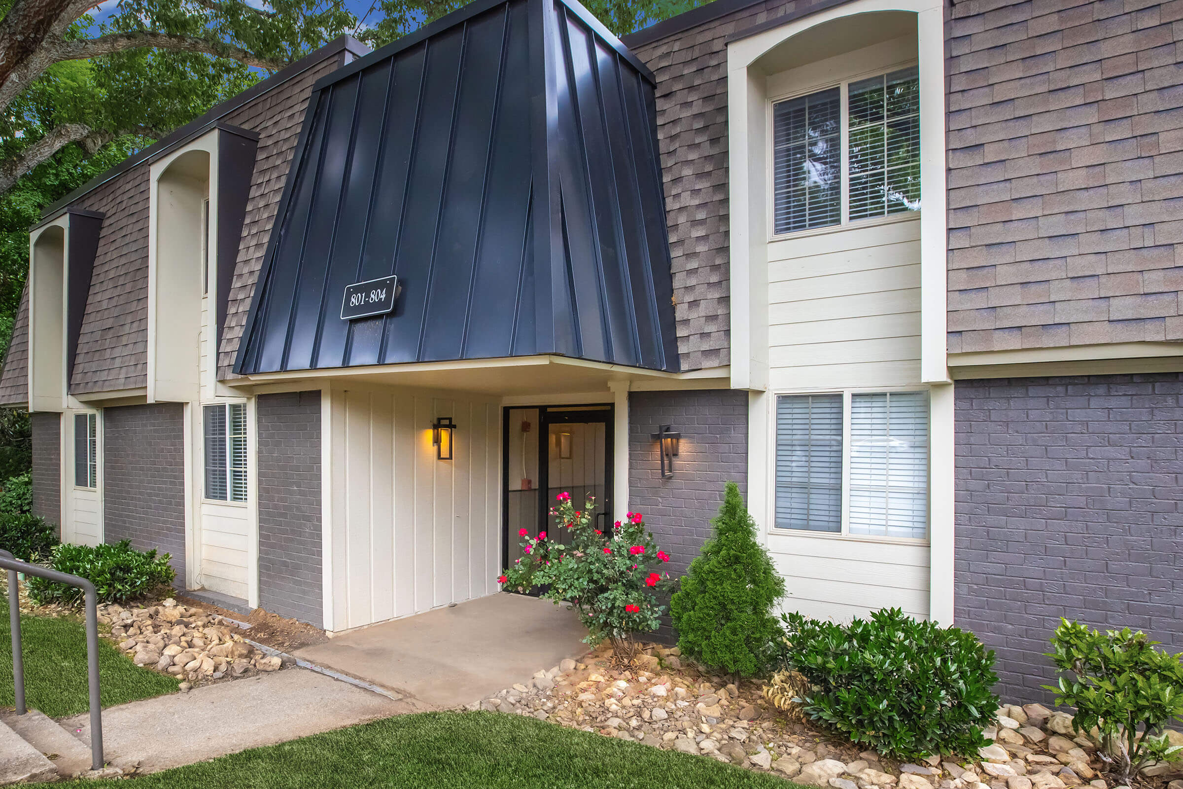 Exterior view of a modern multi-unit apartment building featuring a dark triangular roof, light-colored walls, and well-maintained landscaping. A stone path leads to the entrance, adorned with vibrant rose bushes and small shrubs on either side. Windows allow natural light into the units.