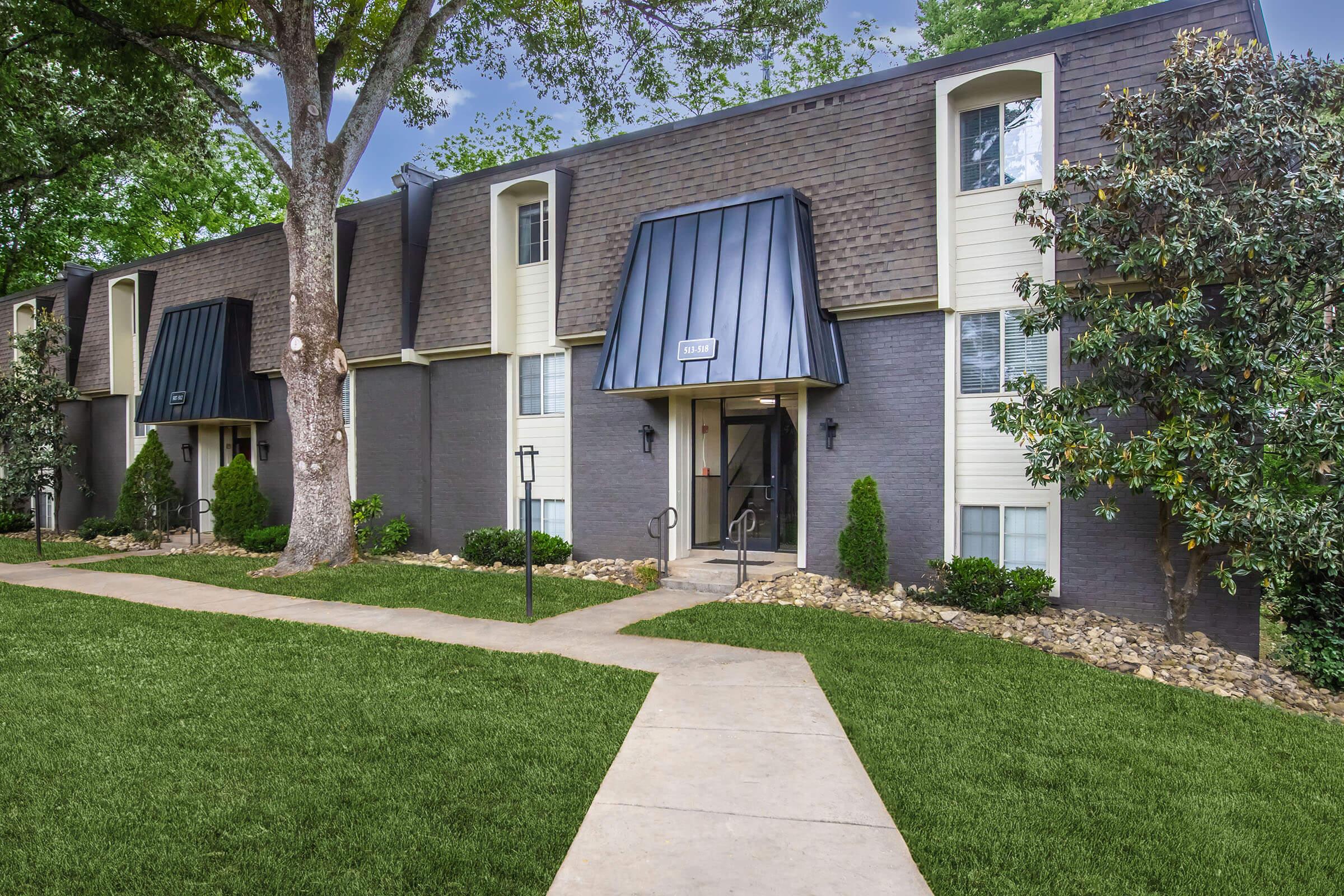 A two-story apartment building with a dark exterior and sloped metal roofs, surrounded by well-manicured lawns and landscaping. A concrete pathway leads to the entrance, flanked by small shrubs and trees. The setting is bright and sunny, suggesting a pleasant residential environment.