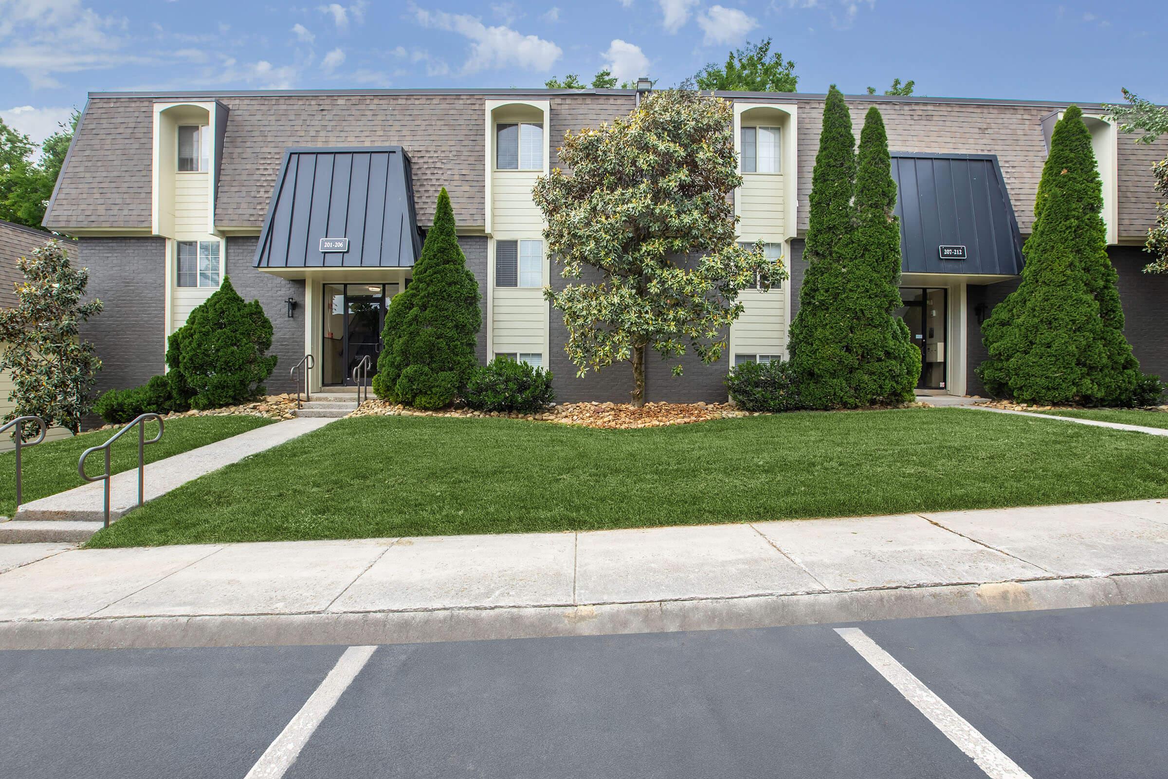 A well-maintained multi-unit residential building with two entrances, surrounded by well-manicured landscaping. The exterior features a combination of dark and light siding, a gray roof, and neatly trimmed shrubs and trees, offering an inviting look. A clear blue sky is visible in the background.