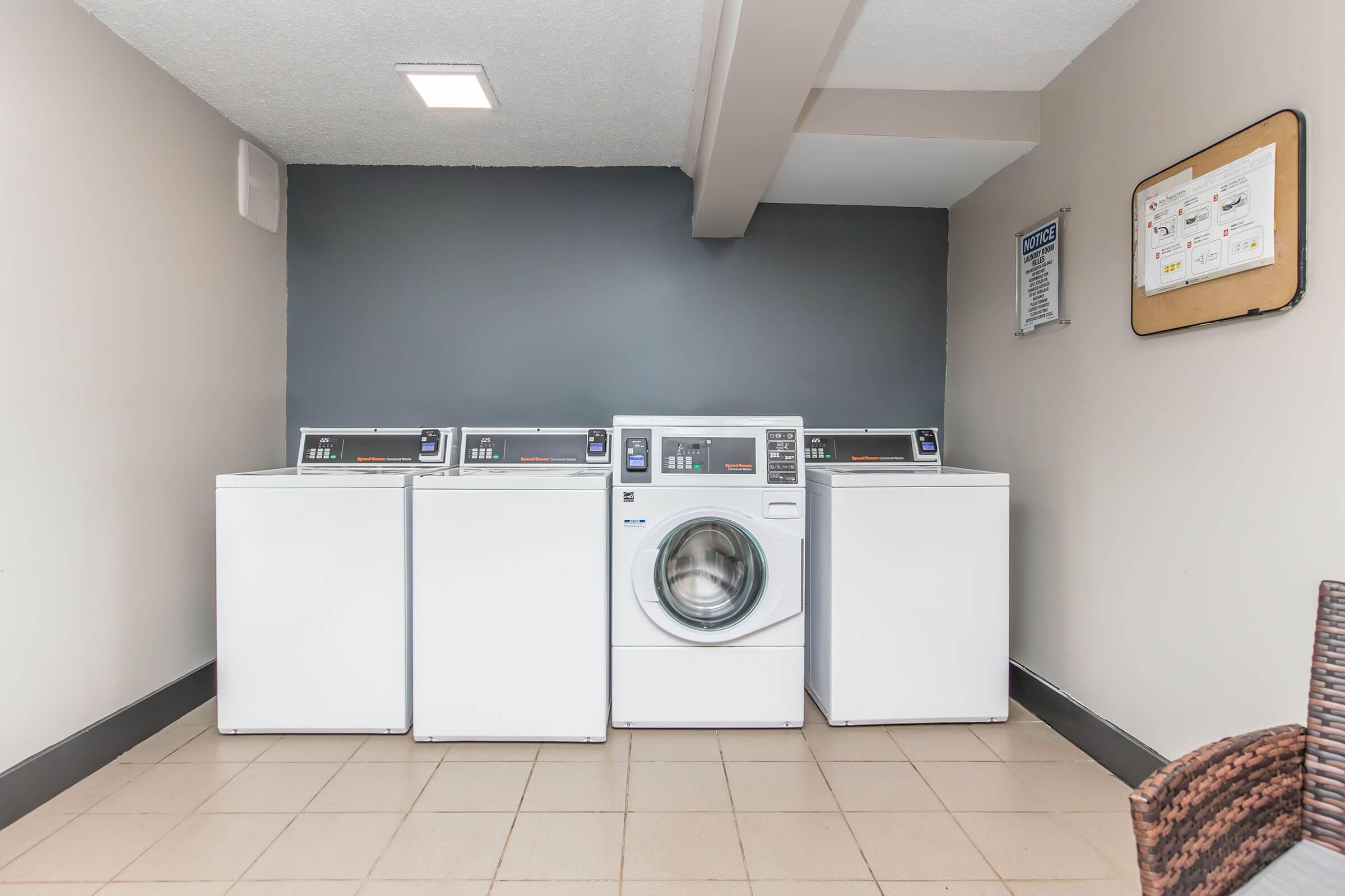 A clean, modern laundry room featuring three front-loading washing machines and one front-loading dryer. The machines are white, positioned against a gray wall, with a bulletin board on the side and tile flooring. Good lighting is evident in the space.