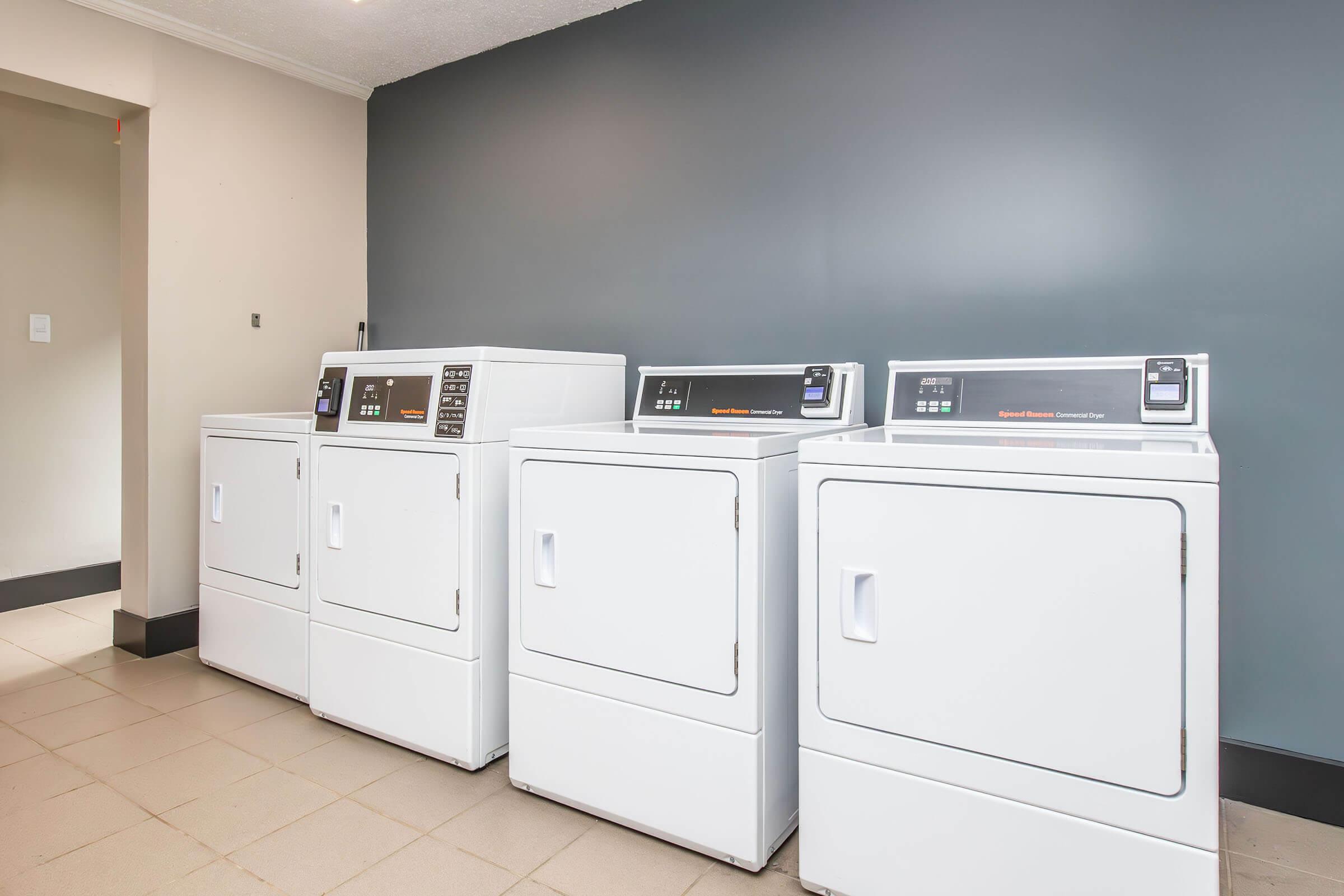 Three white laundry machines, including two dryers and one washer, are lined up against a gray wall in a modern laundry room. The machines have digital displays and buttons for operation. The flooring is tiled in a neutral color.