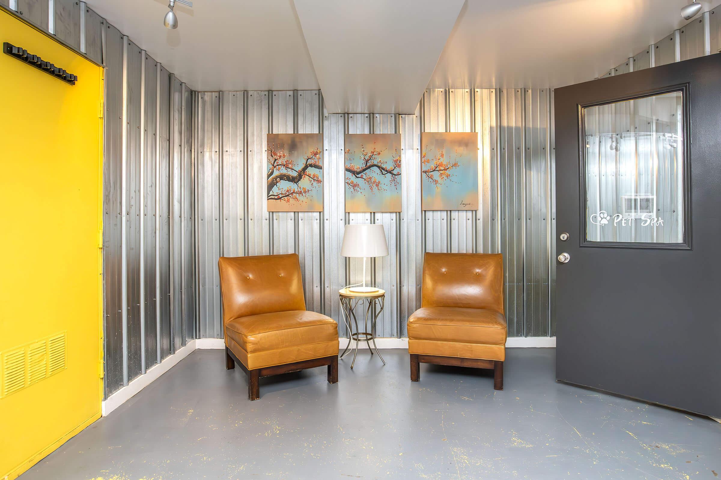 A cozy waiting area featuring two brown leather chairs, a small side table with a lamp, and two framed artworks on the wall. The walls have a metallic finish, with one side painted bright yellow and a door with a glass panel labeled "Pet Spa" visible.