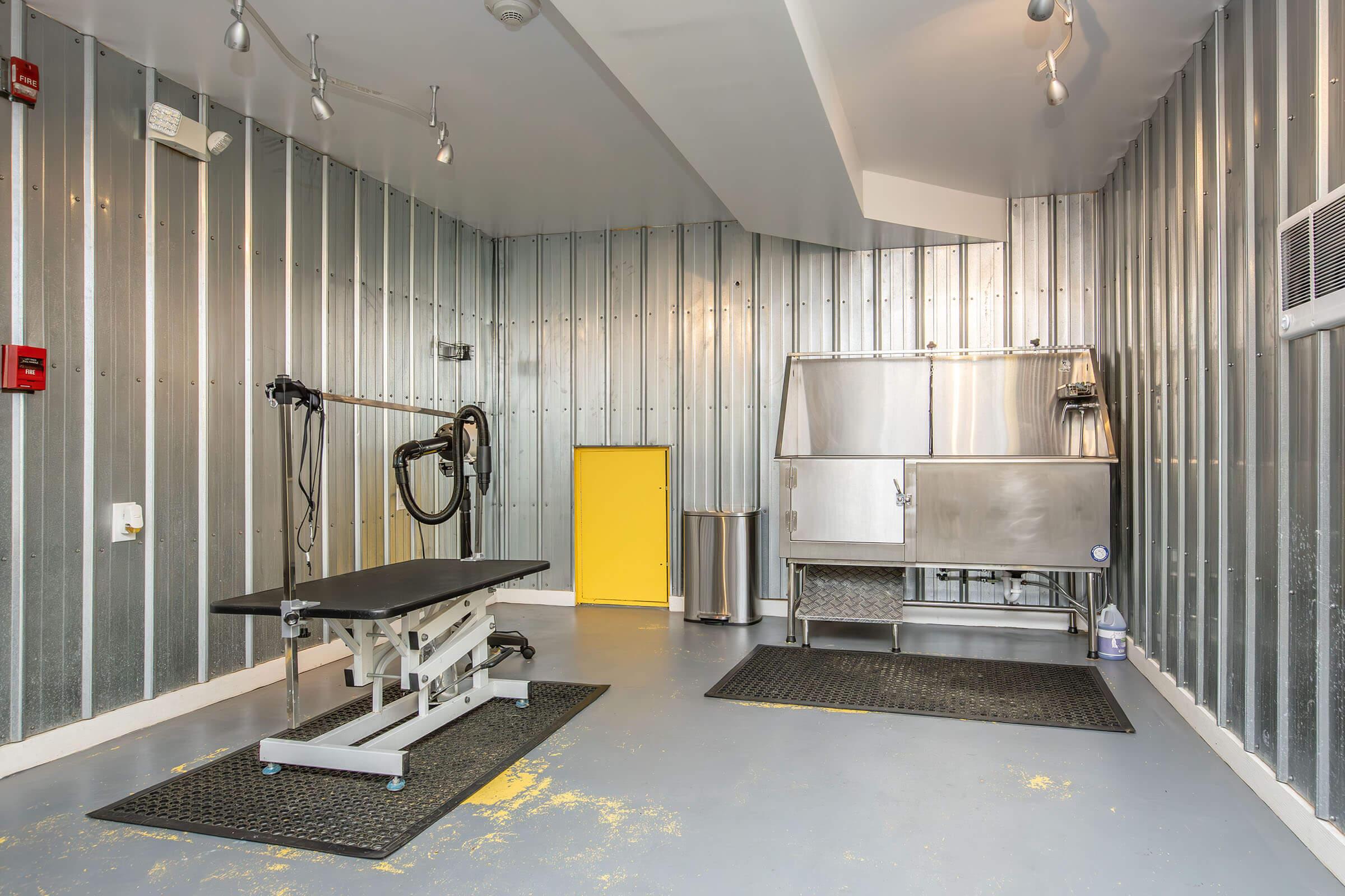 A well-lit, modern grooming room featuring metal walls and gray flooring. It includes a black grooming table with a leash, a stainless steel wash station, and a yellow door. The space is equipped for pet grooming, with mats on the floor for safety and cleanliness.