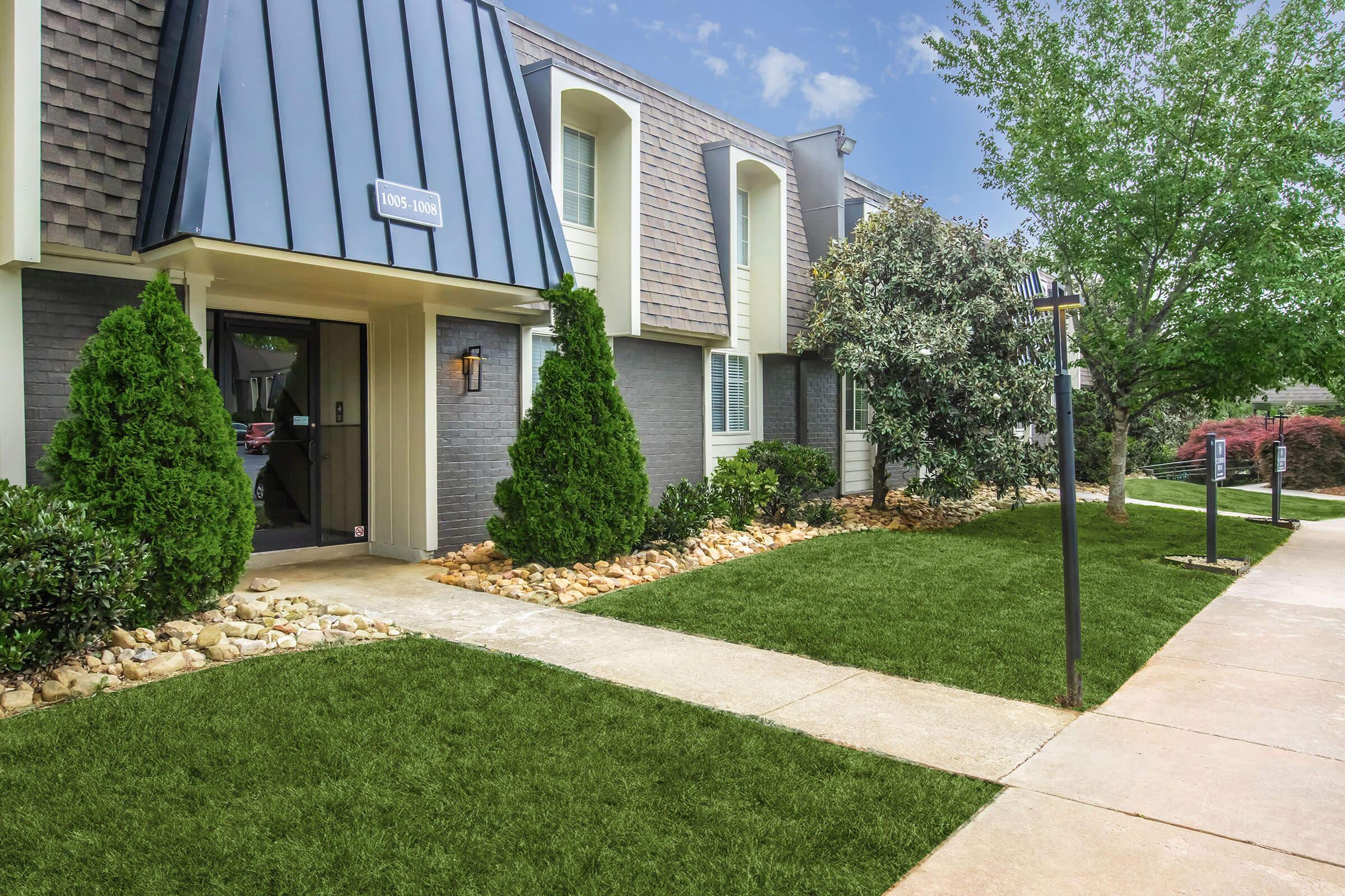 A well-maintained apartment complex featuring a grey exterior with a modern roof design. The entrance has a light fixture and is flanked by manicured green shrubs and rocks. A neatly trimmed lawn and paved walkway lead to the entrance, surrounded by trees and landscaping, creating a welcoming atmosphere.