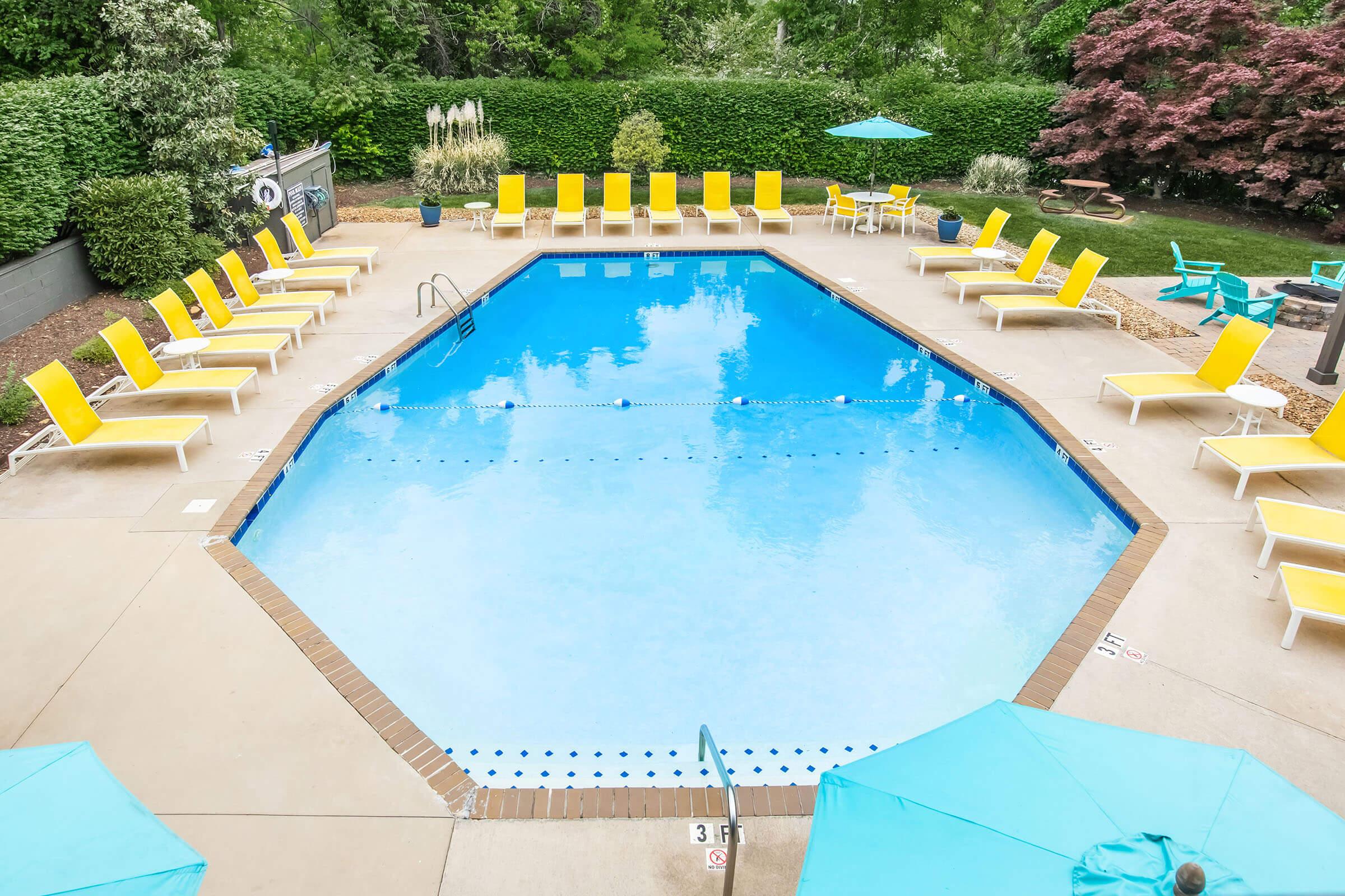 A spacious outdoor pool surrounded by bright yellow lounge chairs, with teal umbrellas providing shade. The pool area is landscaped with greenery and a few trees, creating a relaxing atmosphere. The water is clear and inviting, and the entire scene is well-maintained and inviting for leisure activities.