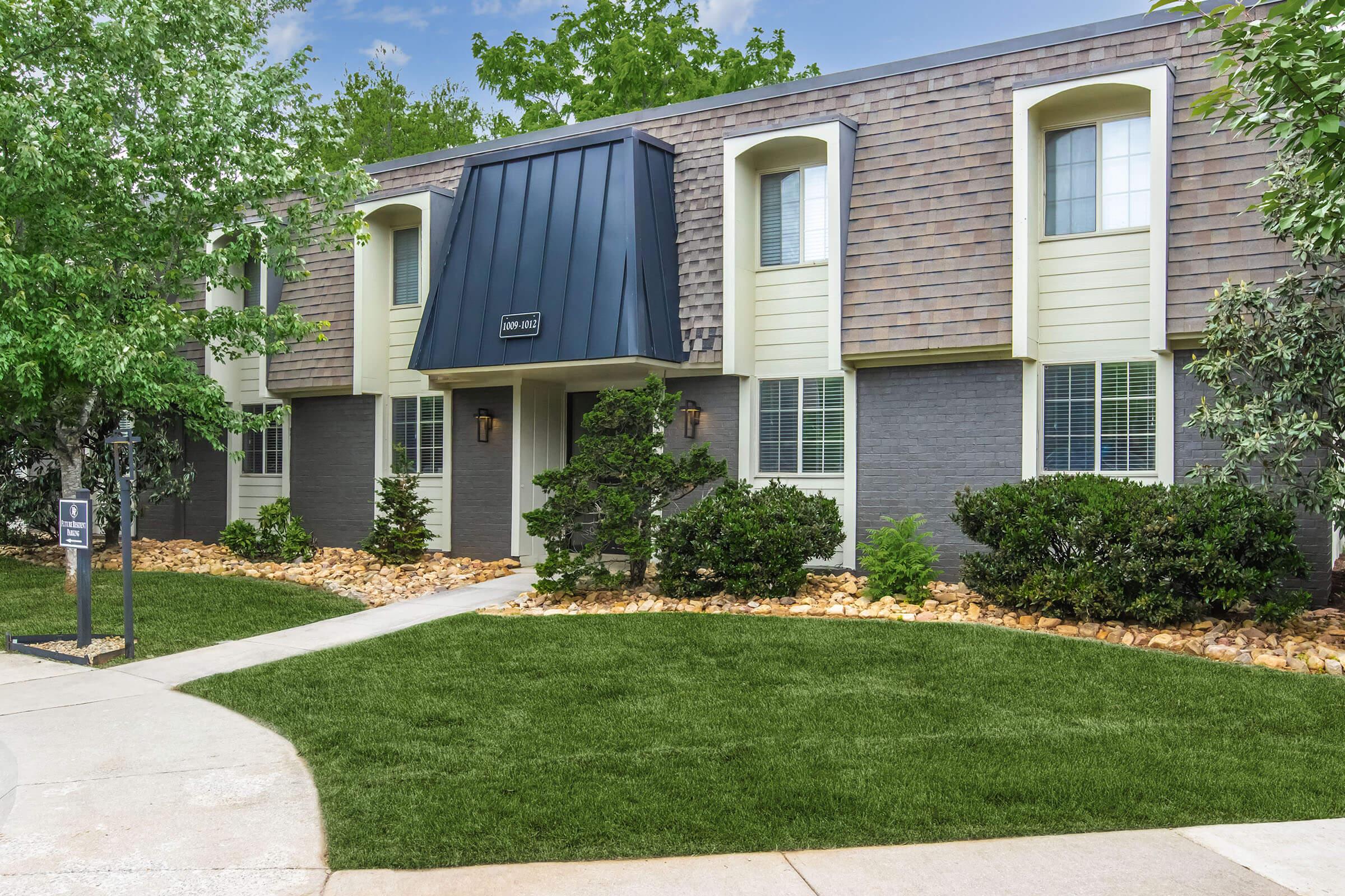 Exterior view of a modern apartment building featuring a gray and beige facade, a distinctive black roof over the entrance, landscaped areas with shrubs and rocks, and a well-maintained lawn. Pathway leading to the entrance, with a sign visible on the left. Clear blue sky and greenery in the background.