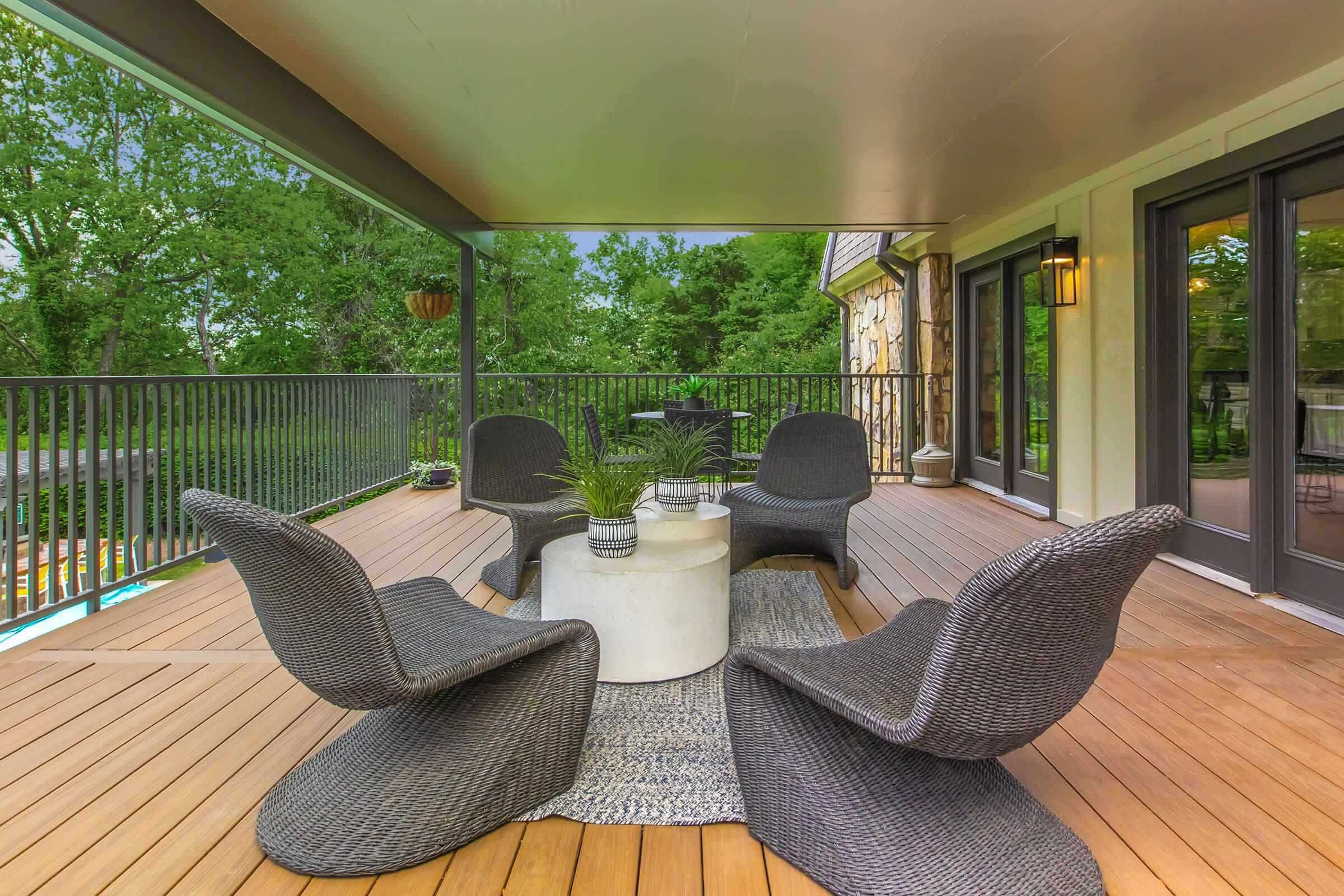 A cozy outdoor seating area featuring four stylish wicker chairs arranged around a round concrete table, set on a wooden deck. A small plant decorates the table, with lush greenery visible in the background, creating a serene atmosphere for relaxation or gatherings.
