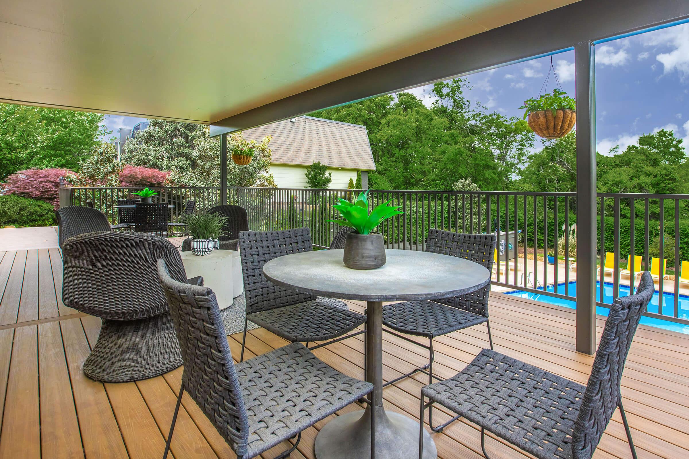 A modern outdoor patio featuring a round table with a potted plant in the center. Surrounding the table are woven chairs. In the background, a lush green landscape and a swimming pool are visible, along with a house peeking through trees. The scene is bright and inviting, perfect for relaxation.