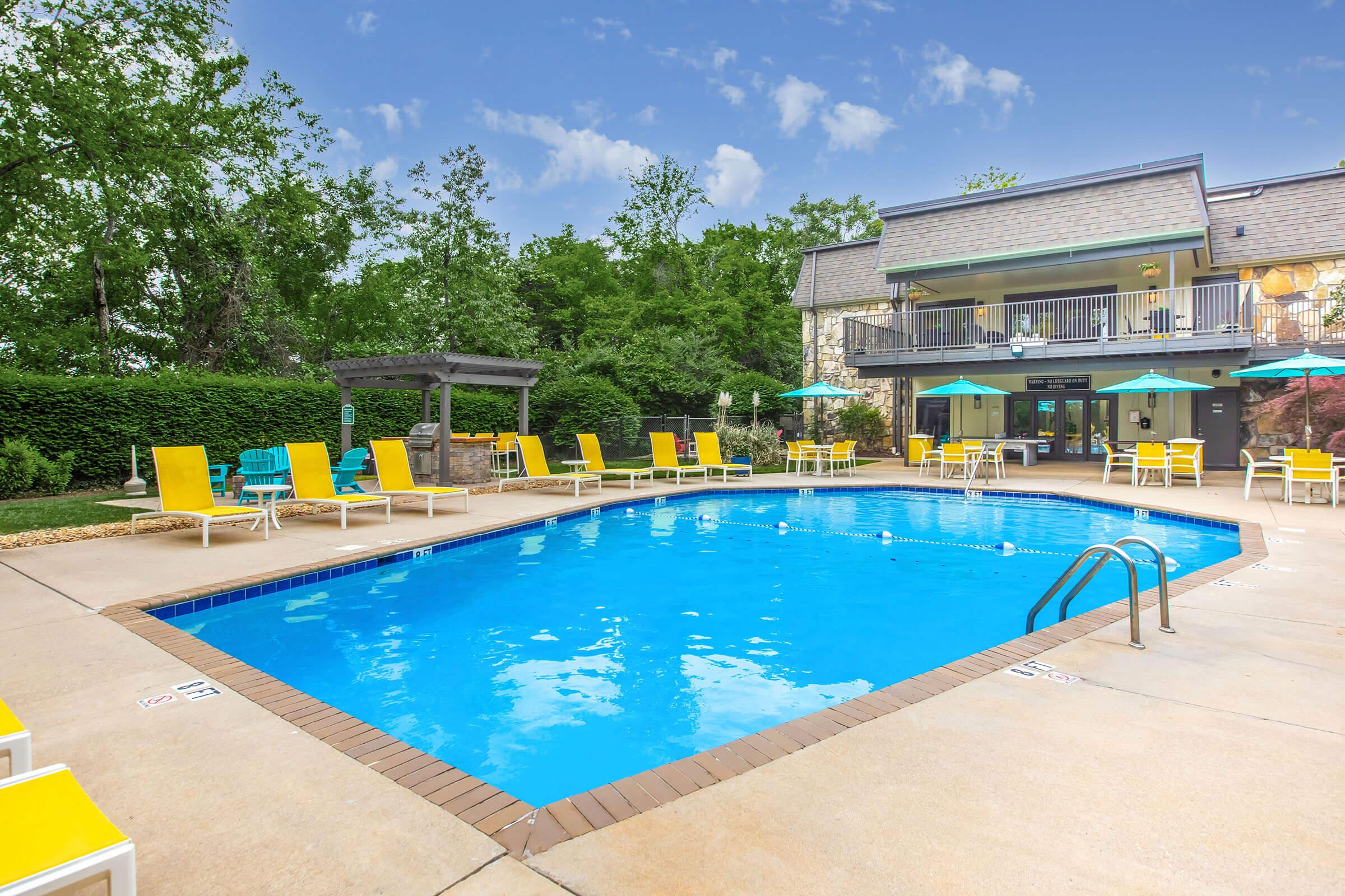 A sunny pool area with clear blue water surrounded by lounge chairs and umbrellas. In the background, a building with multiple balconies and green trees creates a relaxing atmosphere. The scene suggests a tranquil outdoor space ideal for leisure and recreation.