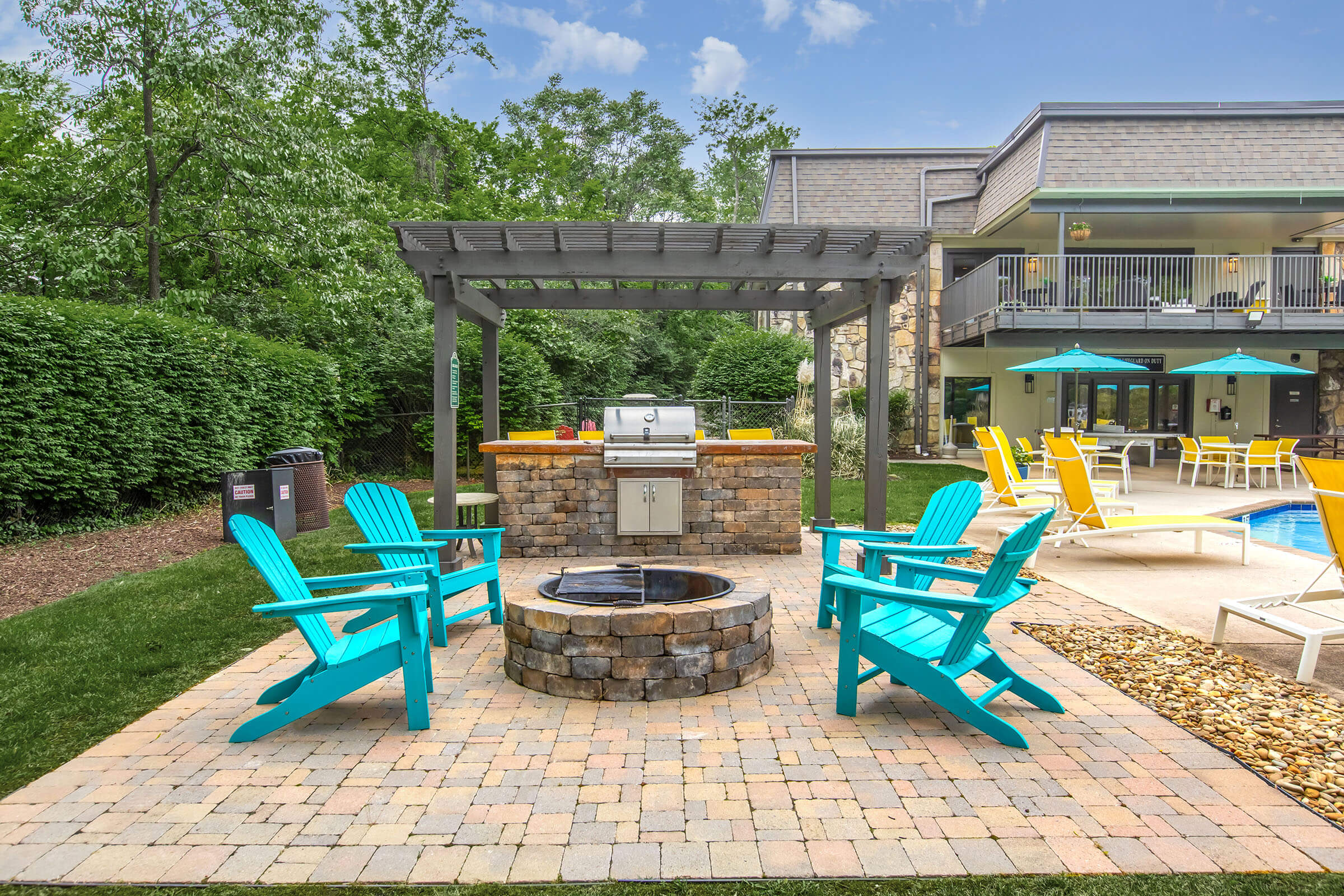 A stylish outdoor seating area featuring bright turquoise Adirondack chairs arranged around a stone fire pit, with a grill under a pergola. The background includes a well-maintained pool area and lounge chairs, surrounded by lush greenery and a clear blue sky.