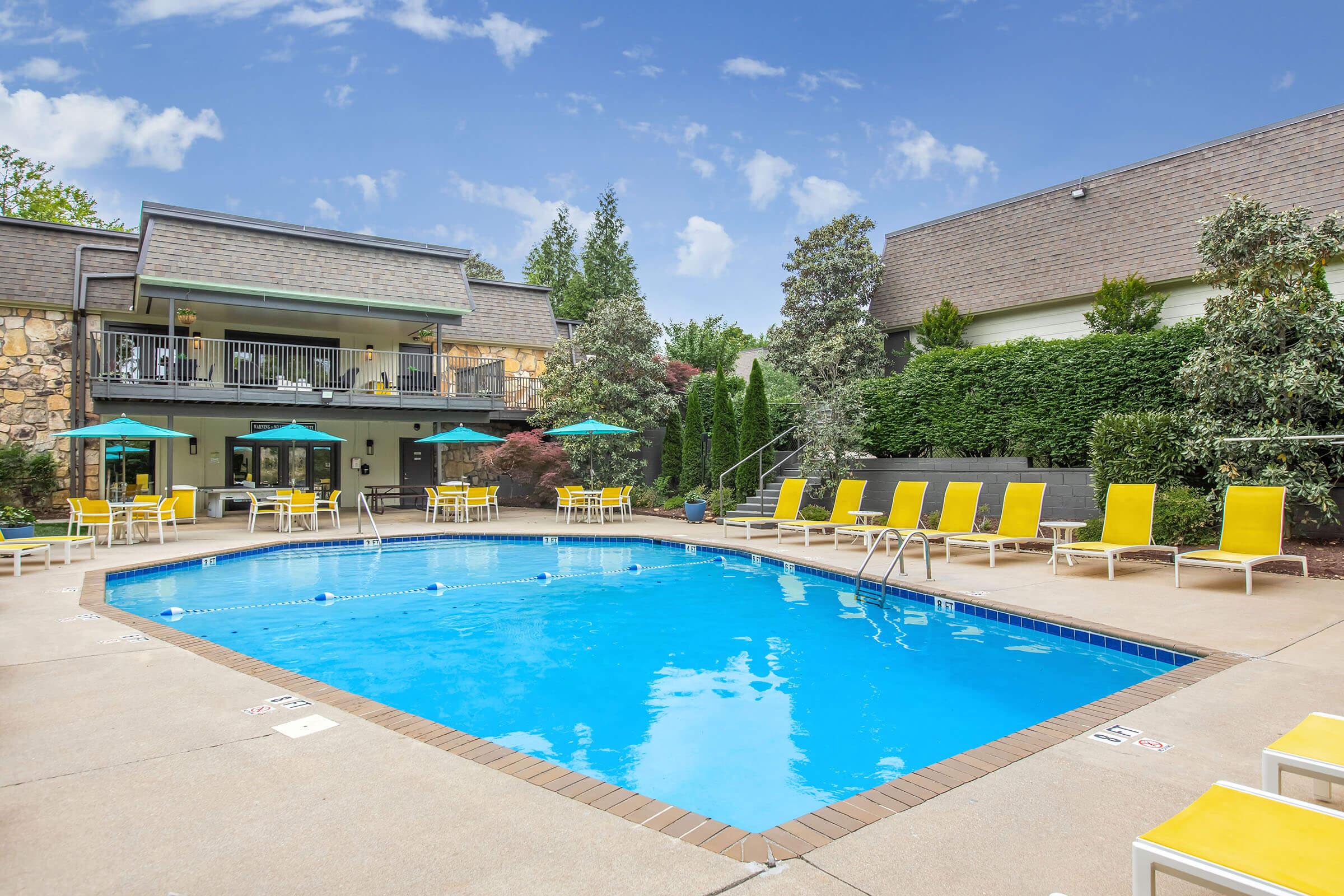 A sunny outdoor swimming pool surrounded by yellow lounge chairs and green umbrellas. Lush landscaping and a building with large windows are visible in the background, creating a relaxing atmosphere ideal for leisure and recreation. The clear blue water reflects the sunny sky above.