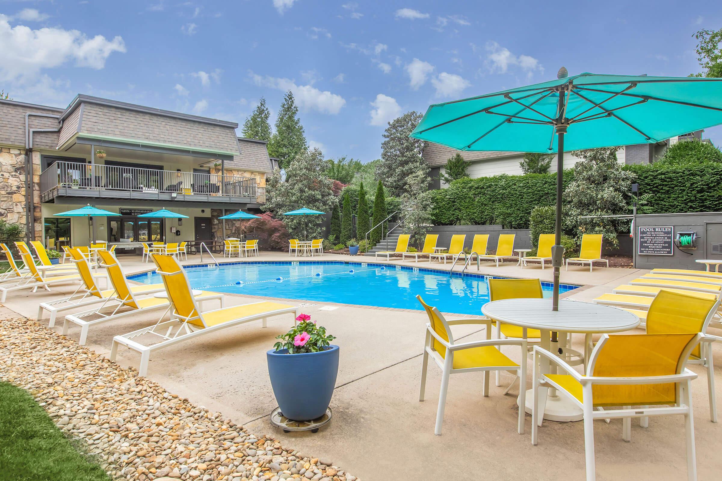 An inviting outdoor pool area featuring a clear blue swimming pool surrounded by bright yellow lounge chairs and tables. Large teal umbrellas provide shade. Lush greenery and decorative flowers enhance the serene atmosphere, with a modern building visible in the background under a partly cloudy sky.