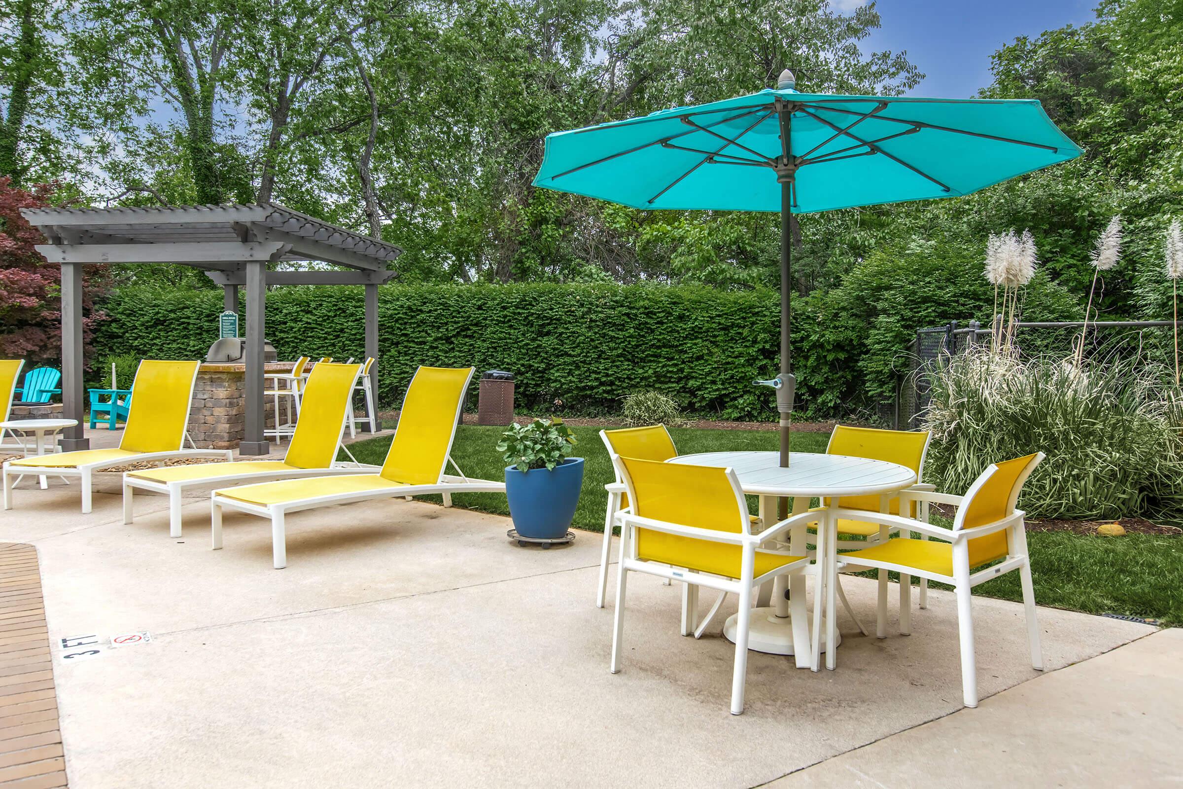 A bright outdoor patio scene featuring yellow lounge chairs, a small dining table with yellow chairs, and a large turquoise umbrella. Lush green trees and shrubs provide a natural backdrop, while a blue planter adds a pop of color to the setting. The area is paved, creating a cozy, inviting atmosphere for relaxation.