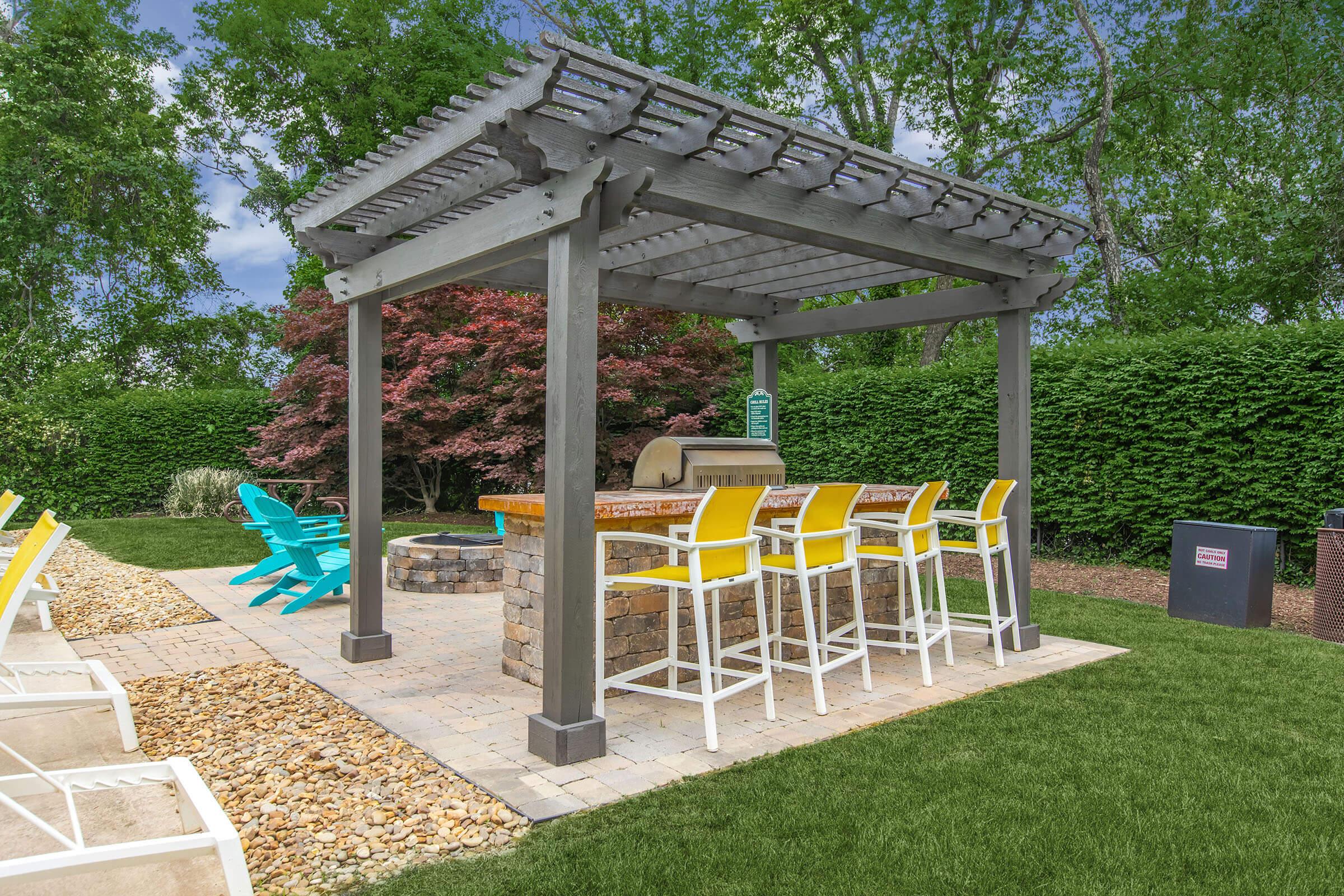 An outdoor bar area with a wooden pergola, featuring high stools in vibrant yellow. The bar top is made of stone, and there's a fire pit nearby. Surrounding greenery adds to the serene atmosphere, with lounge chairs in the foreground and well-maintained grass. It's a perfect setting for relaxation and social gatherings.