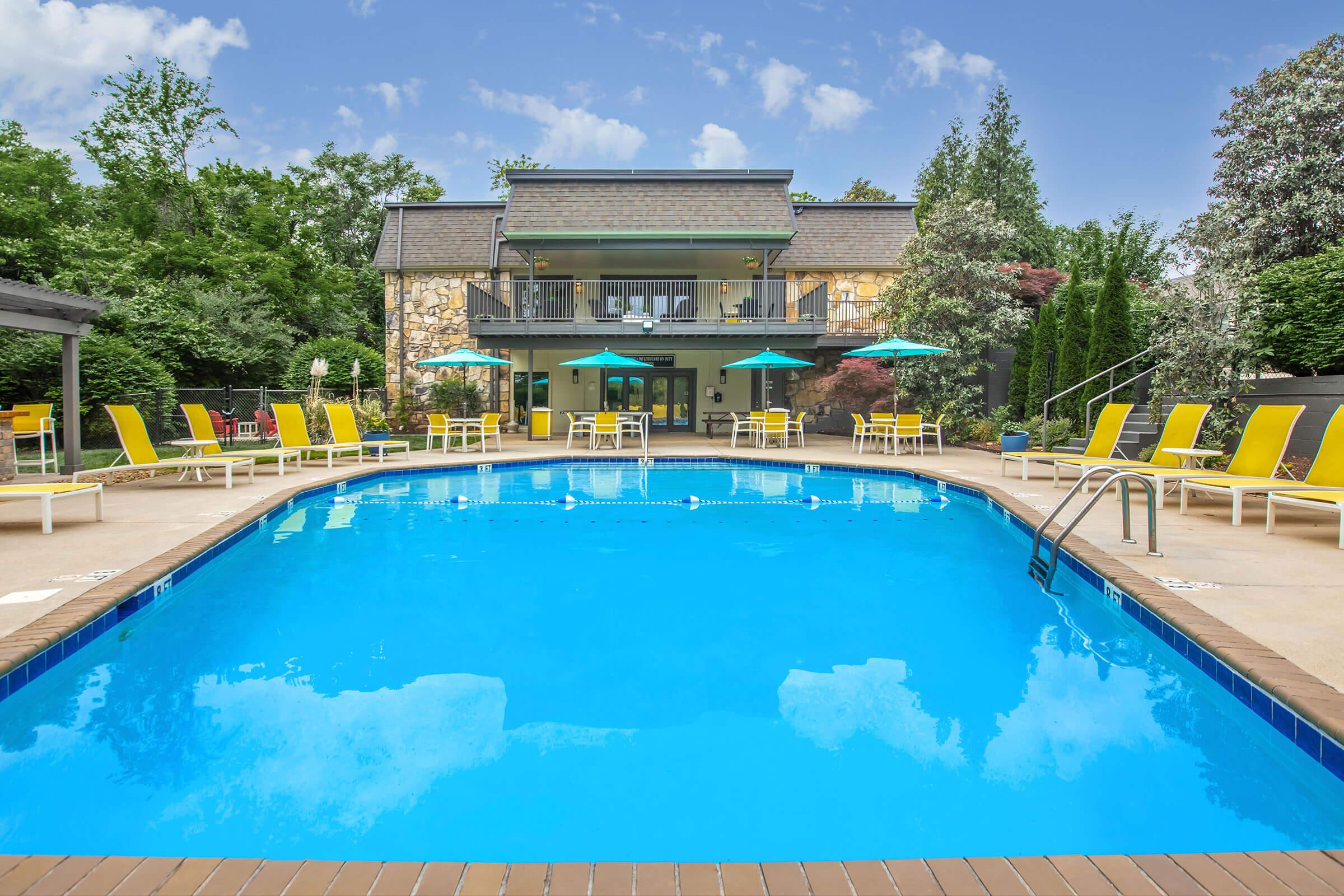 A clear blue swimming pool surrounded by yellow lounge chairs and green umbrellas. In the background, a multi-story building with a stone facade and large windows sits among trees and landscaped greenery. A sunny sky with scattered clouds completes the serene setting.