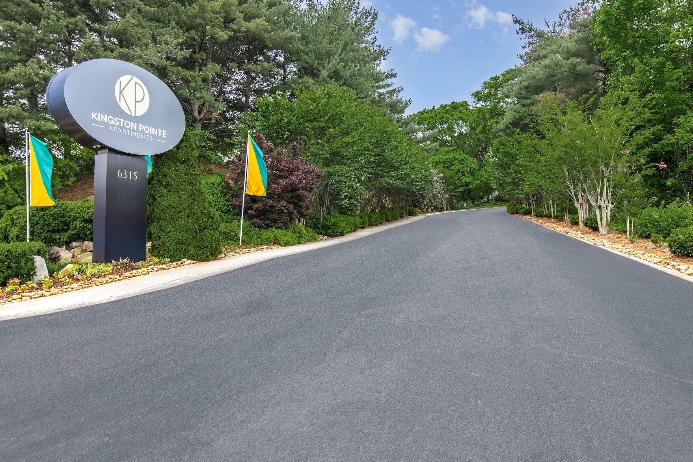 A well-maintained asphalt road leading to Kingston Pointe Apartments, flanked by decorative signage on the left. Lush greenery and colorful flags are visible along the sides, creating an inviting atmosphere. The scene is bright and sunny, with clear skies and a vibrant landscape.