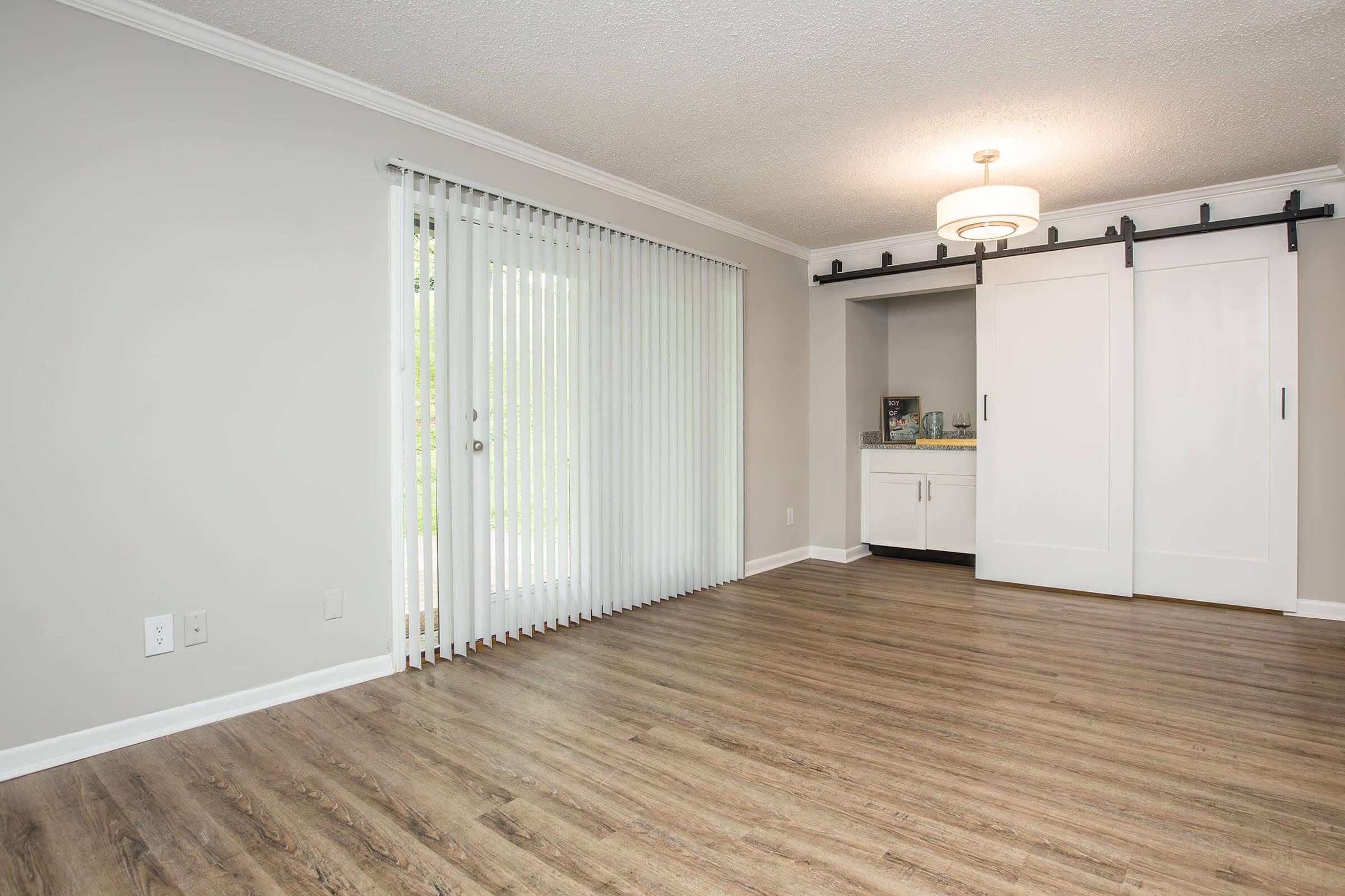 A spacious, well-lit room with light gray walls and wooden flooring. It features a set of vertical blinds on a glass door leading to the outdoors, a modern ceiling light fixture, and a closet with sliding white doors. The overall decor is minimalist and inviting.