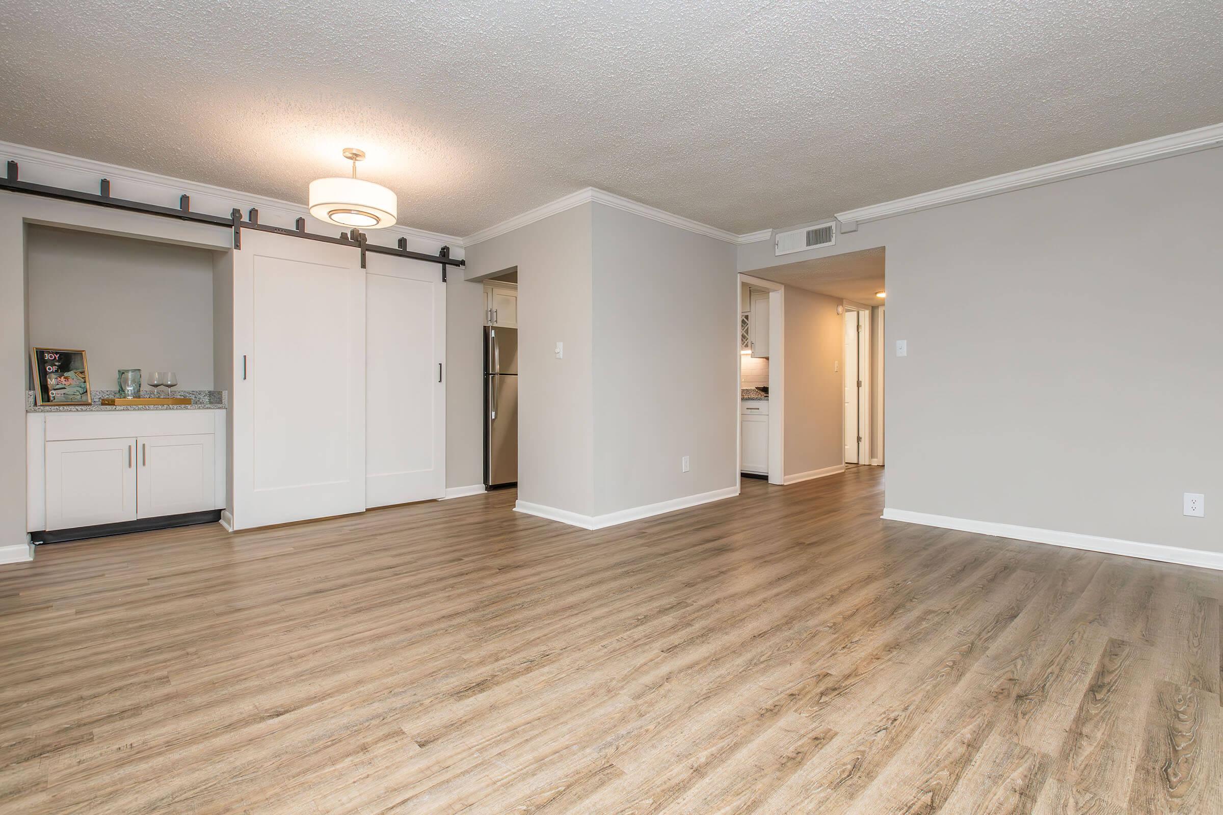 A spacious, well-lit living area featuring light brown wood flooring, neutral-colored walls, and a modern ceiling light fixture. Sliding doors lead to a closet area, while a kitchenette is visible in the background, providing a sense of openness and contemporary design.