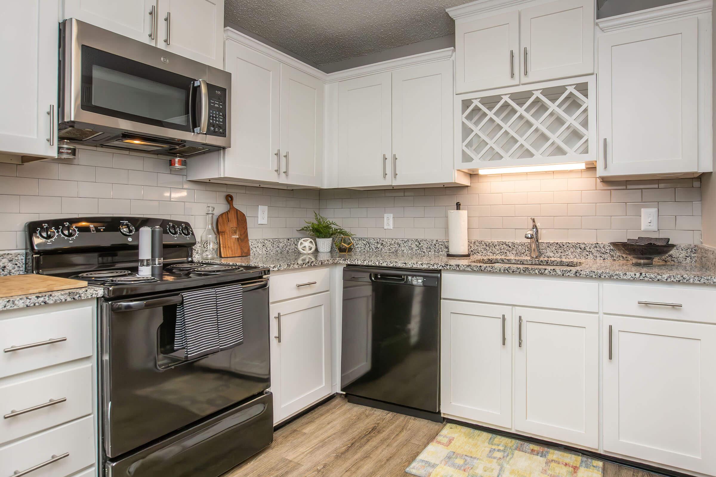 A modern kitchen featuring white cabinetry, a stainless steel microwave, a black stove and dishwasher, and a granite countertop. The backsplash is tiled in light gray, with a decorative wine rack above the sink. A wooden cutting board and potted plants add decorative elements to the space.