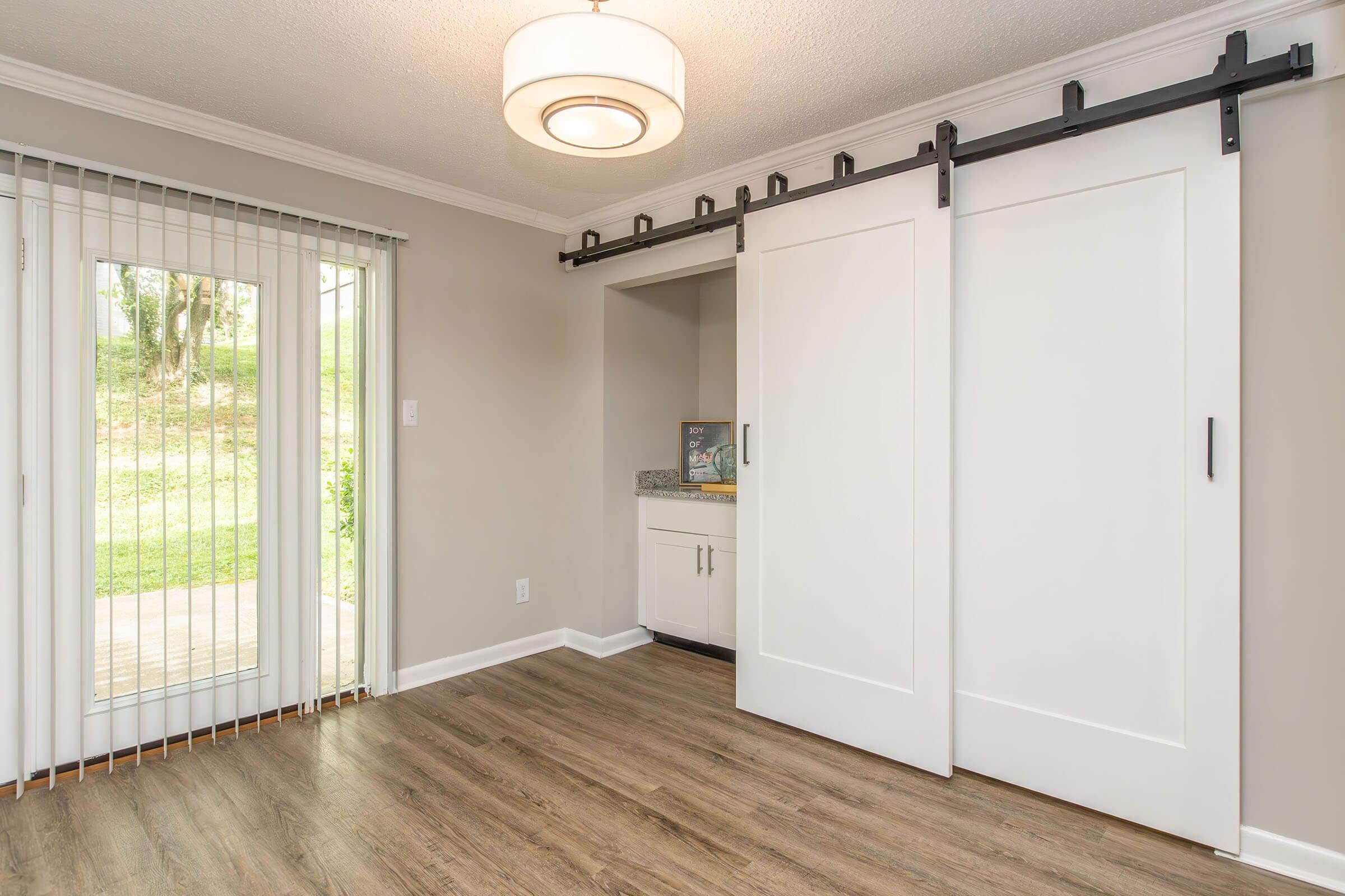 A well-lit interior room featuring beige walls and a wooden floor. There are two large white sliding doors on a black track, leading to a small kitchenette area with a countertop. A sliding glass door opens to an outdoor area, and natural light filters in through the windows, enhancing the spacious feel.