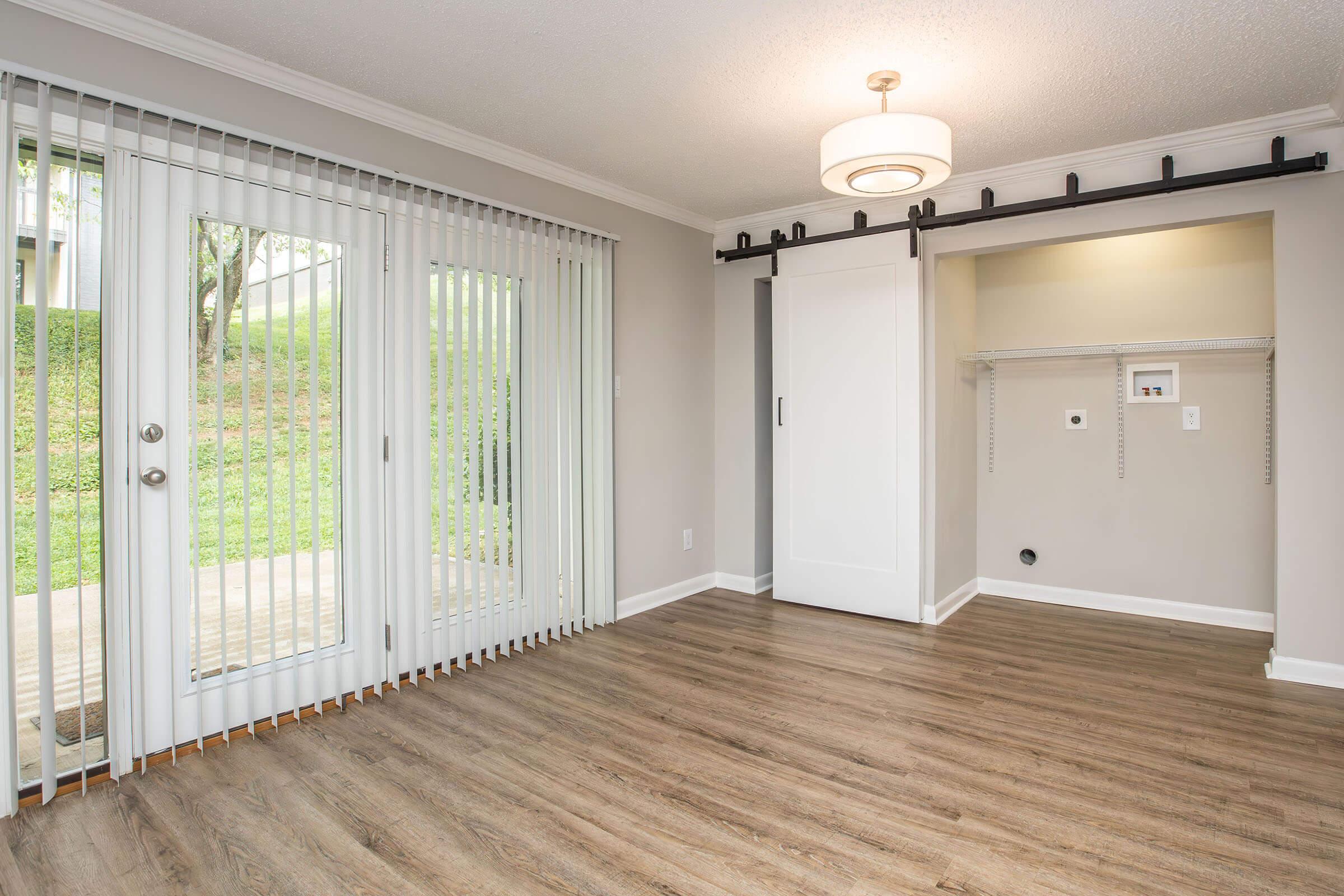 A spacious room with light-colored walls and wooden flooring. It features a sliding glass door with vertical blinds leading to an outdoor area, a modern light fixture, and a white sliding closet door. There is a small nook with shelving and electrical outlets.