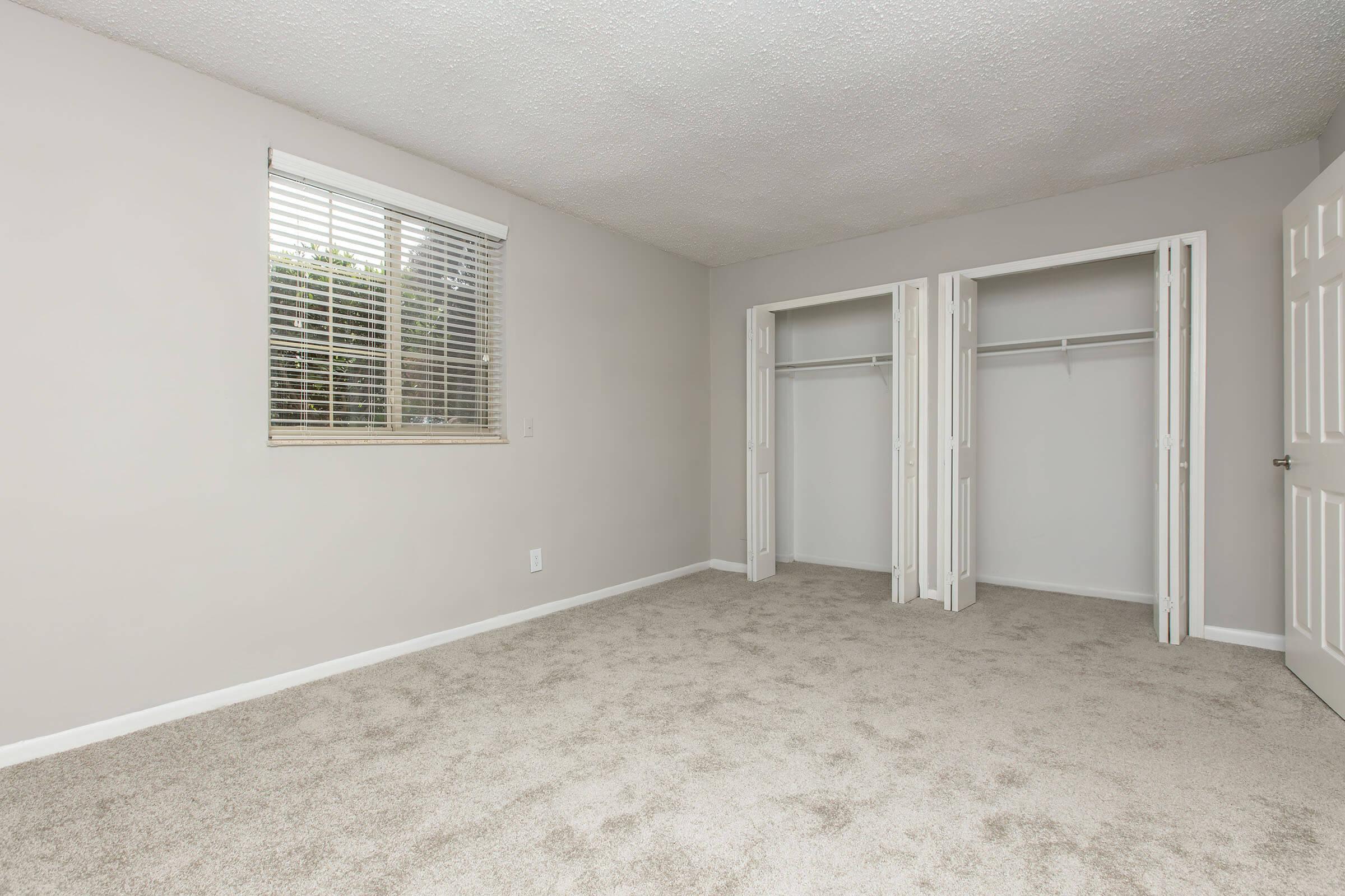 Empty room with light gray walls and a plush carpet. There is a window with horizontal blinds allowing natural light. Two built-in closets with double doors are visible along one wall, and the overall space appears tidy and spacious.