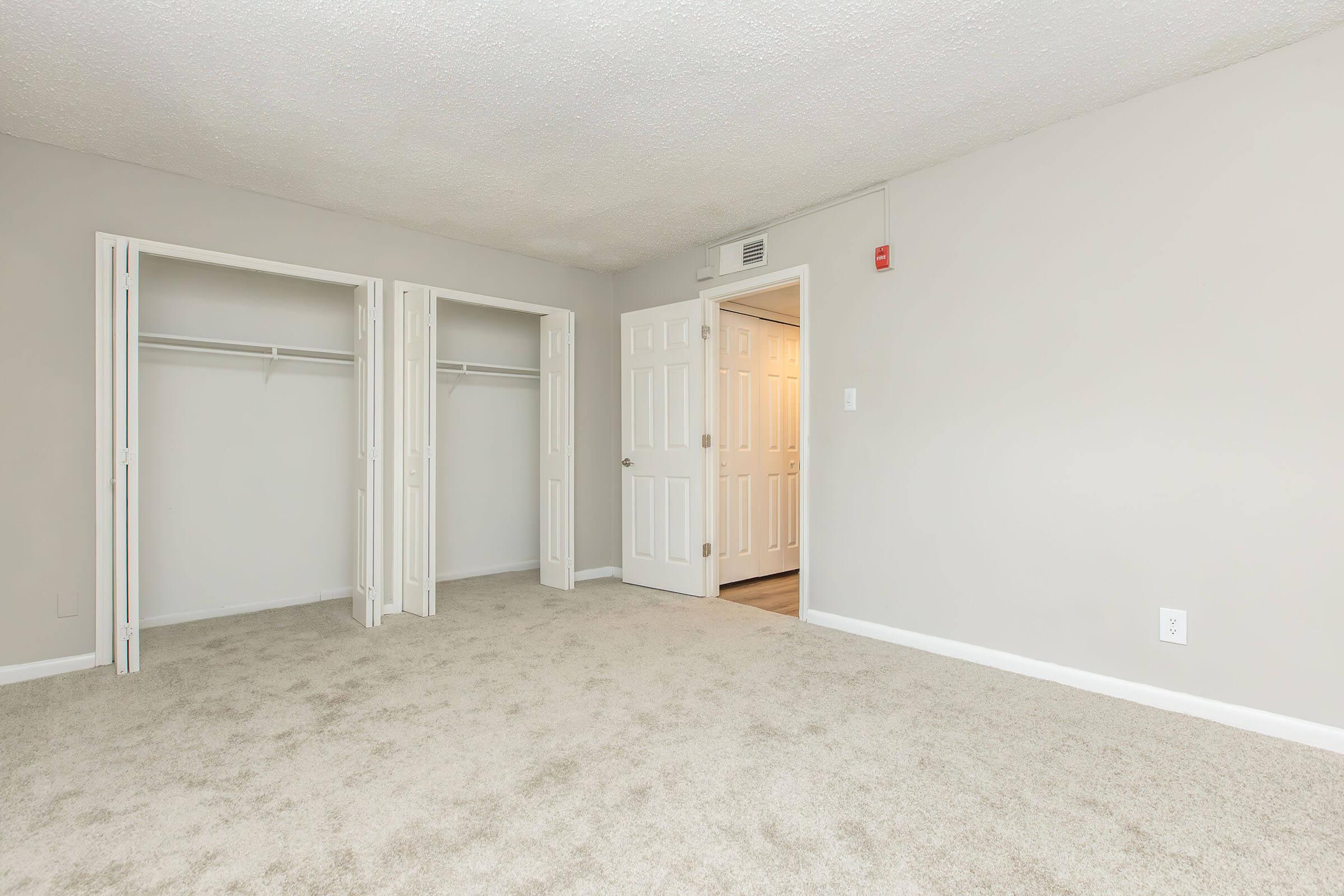 A spacious, empty bedroom featuring light gray walls and soft carpeting. Two closet doors are open on the left, showing ample storage space. A doorway on the right leads to another room, illuminated by soft lighting, creating a bright and inviting atmosphere.