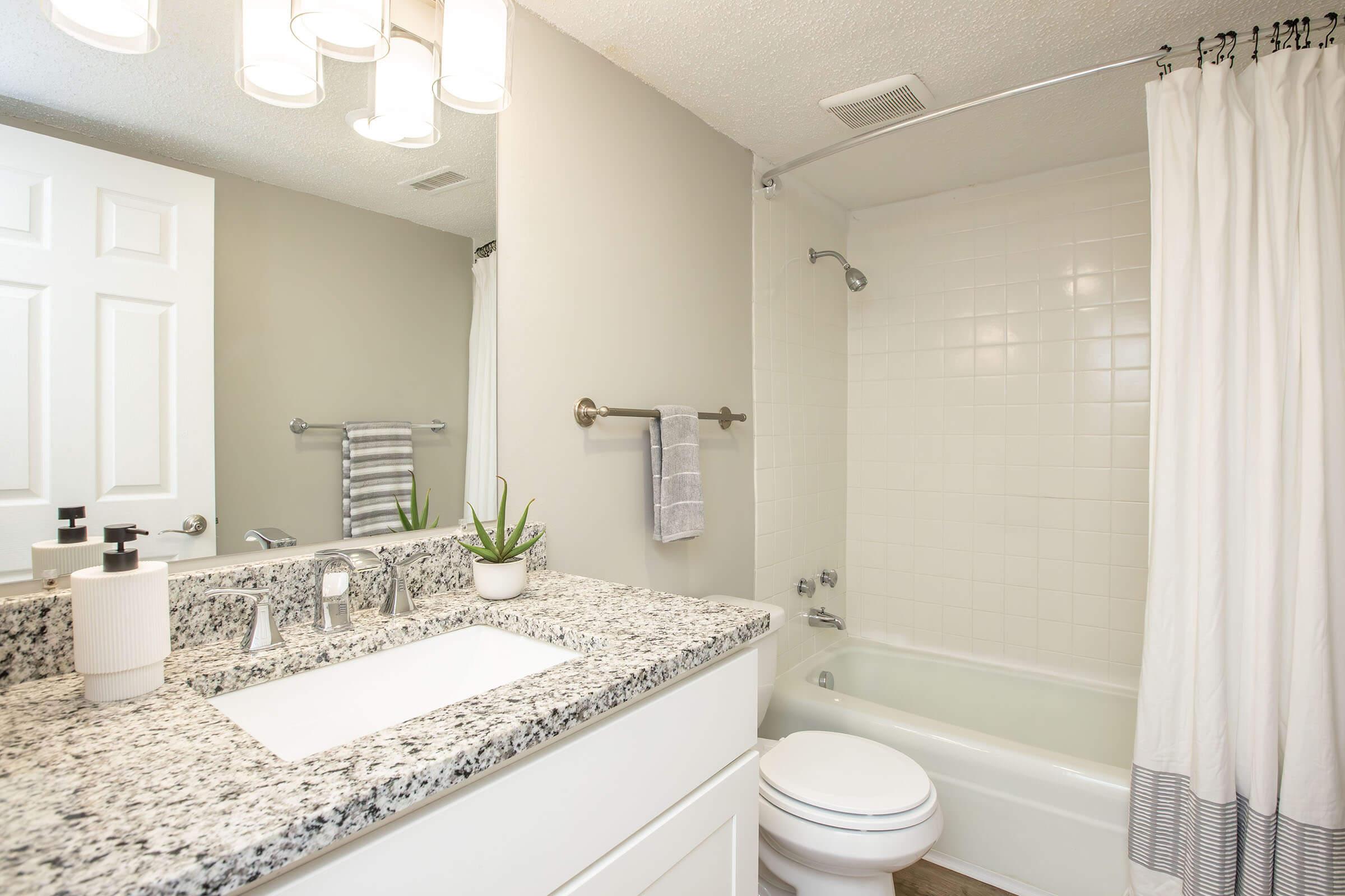 A clean, modern bathroom featuring a white countertop with a sink, a large mirror, and decorative items. The bath area has a white tile tub with a shower curtain. Light fixtures above the mirror provide bright lighting. A towel bar with a gray towel and a small plant add a touch of decor.