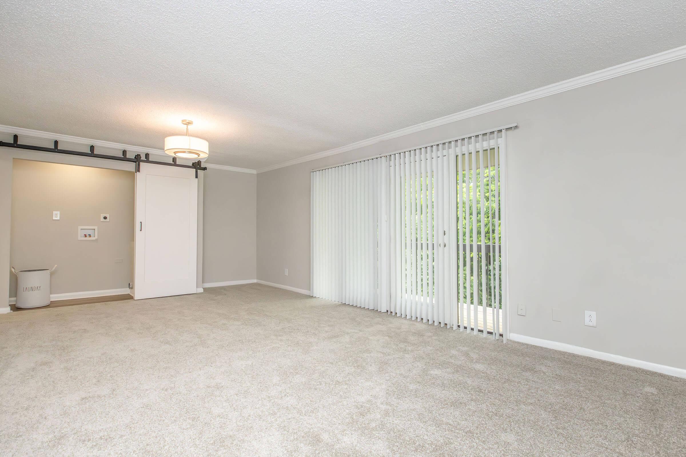 A spacious living room with light beige carpet and neutral-colored walls. There’s a sliding glass door with vertical blinds leading to a balcony, and a modern light fixture hanging from the ceiling. A small area with wall-mounted features and a concealed storage space is visible.