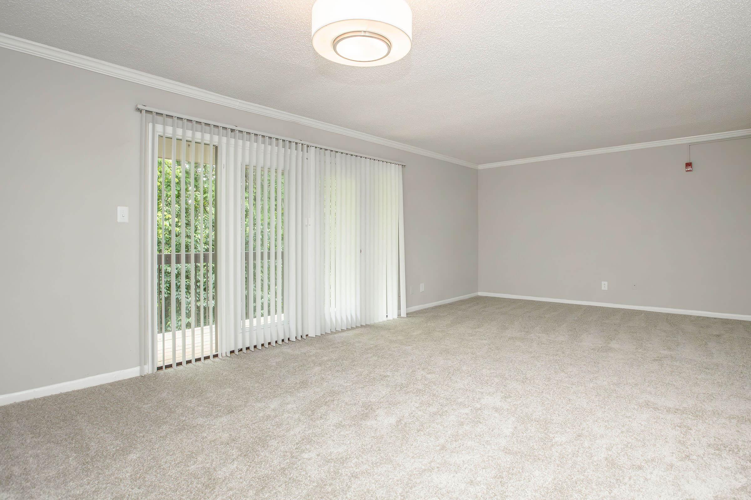 A spacious, empty living room featuring light gray walls and plush beige carpet. Large windows with vertical blinds allow natural light to enter, leading to a balcony outside. A modern circular light fixture hangs from the ceiling, adding a contemporary touch to the room.