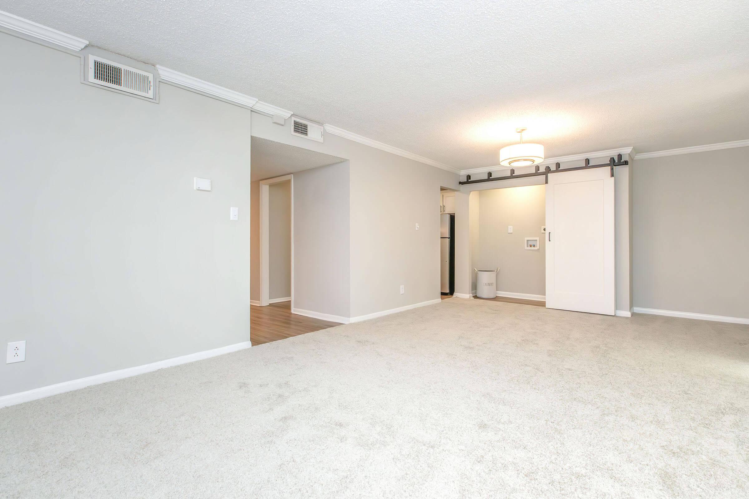 A spacious, empty living room with light gray walls and carpet. There is a white door on the right, leading to another room, and a small kitchen area visible in the background. A ceiling light fixture provides soft illumination, creating a bright, airy atmosphere.
