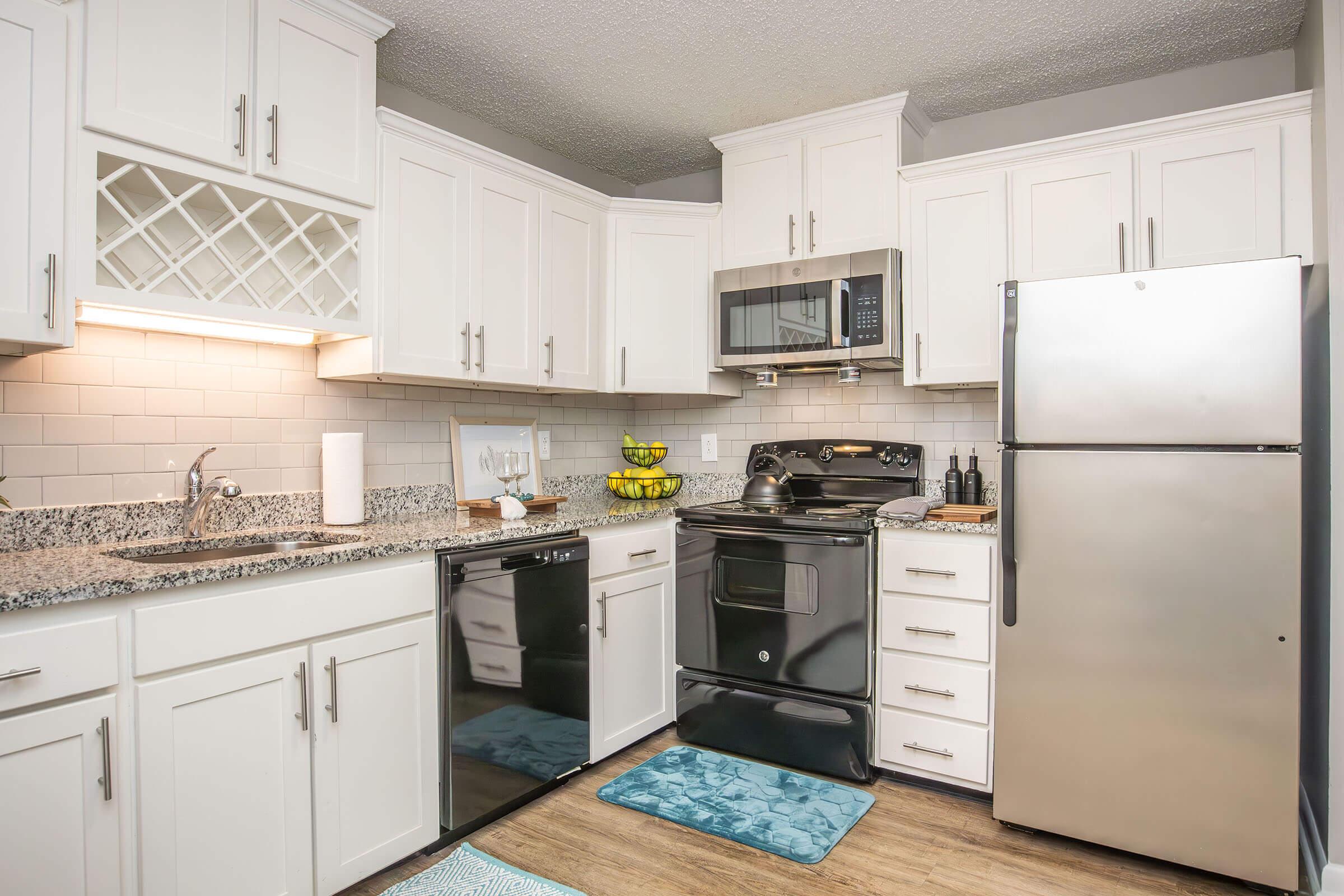 A modern kitchen featuring white cabinetry, a granite countertop, and stainless steel appliances. The setup includes a black stove, microwave, and refrigerator, with a dishwasher under the counter. A bowl of bananas sits on the countertop, and a light blue rug adds a pop of color to the floor.