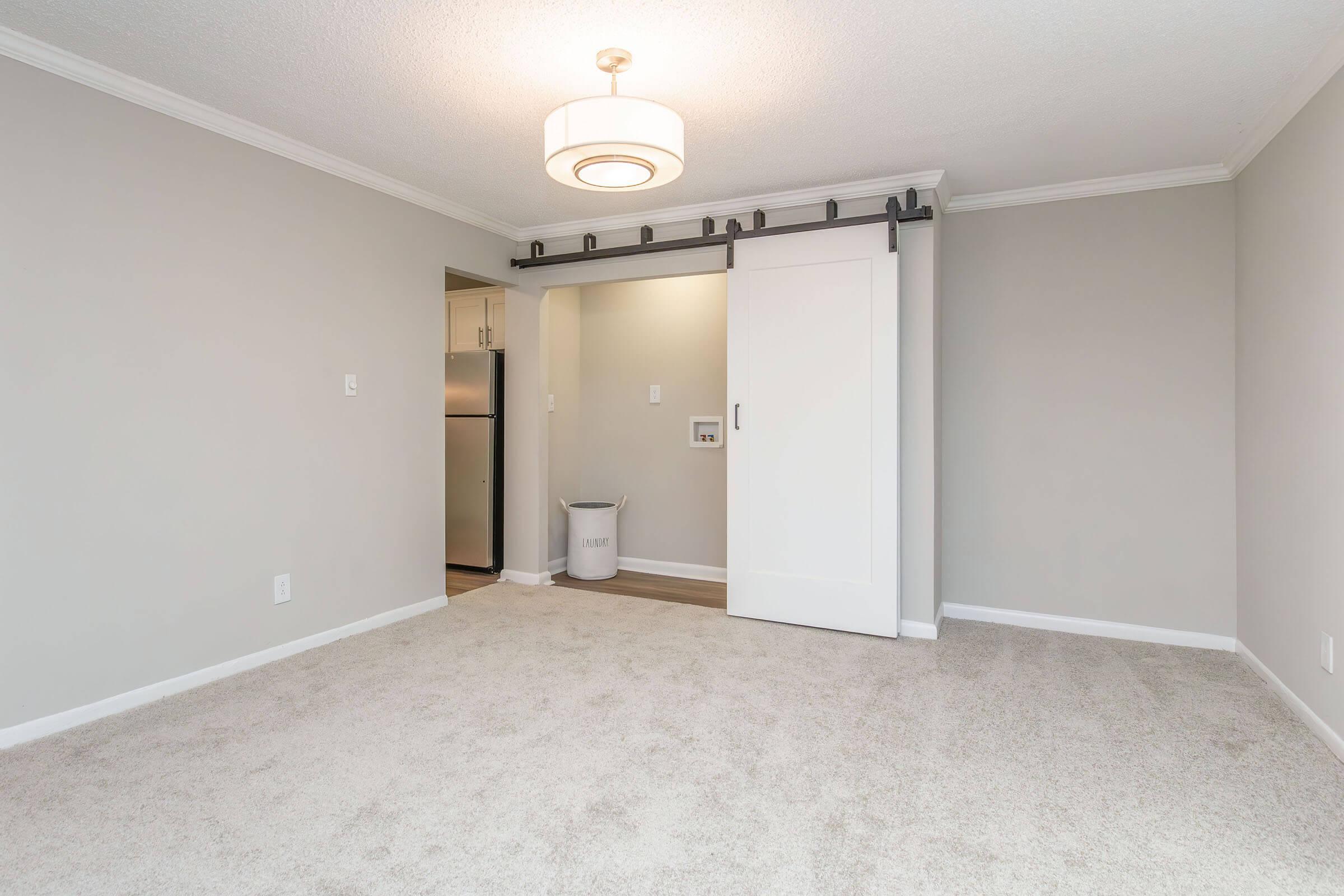 A spacious room with light gray walls and soft carpet. It features a modern ceiling light fixture and a sliding white door leading to a small kitchen area. A waste bin is visible near the door, and there's ample natural light suggesting a bright and airy atmosphere.