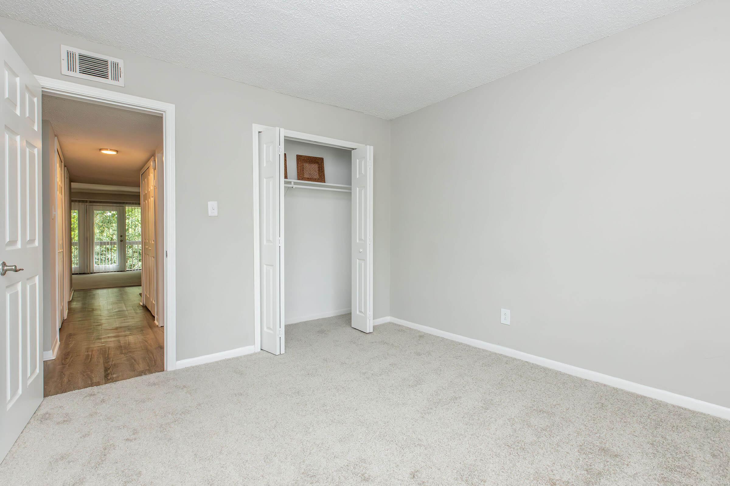 Empty bedroom with beige carpet, neutral gray walls, and a closet with double doors. A doorway leads to a hallway with natural light coming from the adjacent room. The space is minimalistic and well-lit, creating a feeling of openness.