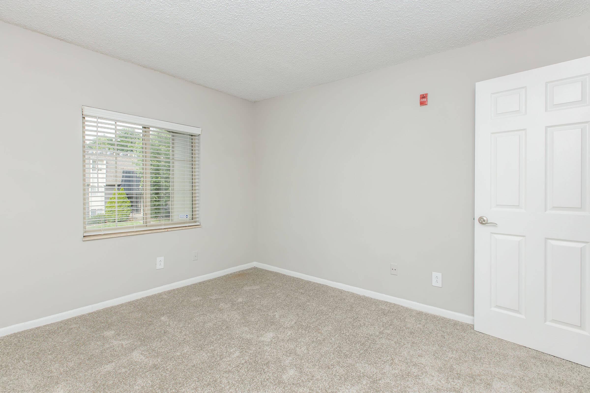 Empty room with light gray walls and beige carpet. A small window with blinds allows natural light in, and a white door is visible on the right side of the image. No furniture or decorations are present, giving the space a clean and minimalist appearance.