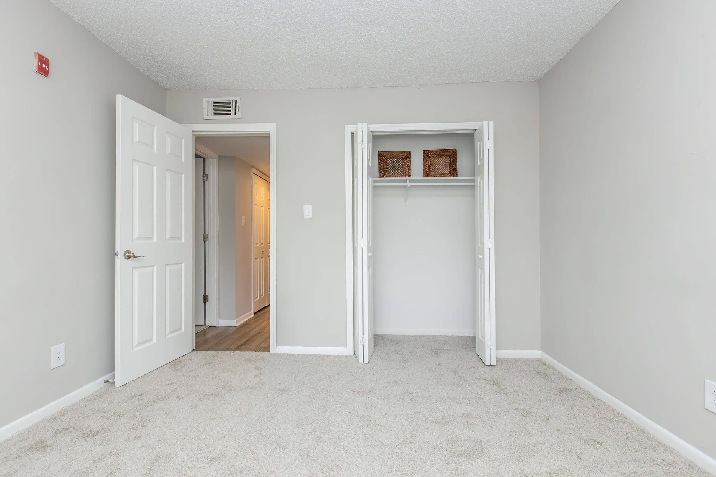 A view of a vacant bedroom with light gray walls and plush carpet. A door on the left leads to another room, while a closet with double doors is on the right, showcasing empty shelves. The space is well-lit with natural light, creating a clean and inviting atmosphere.