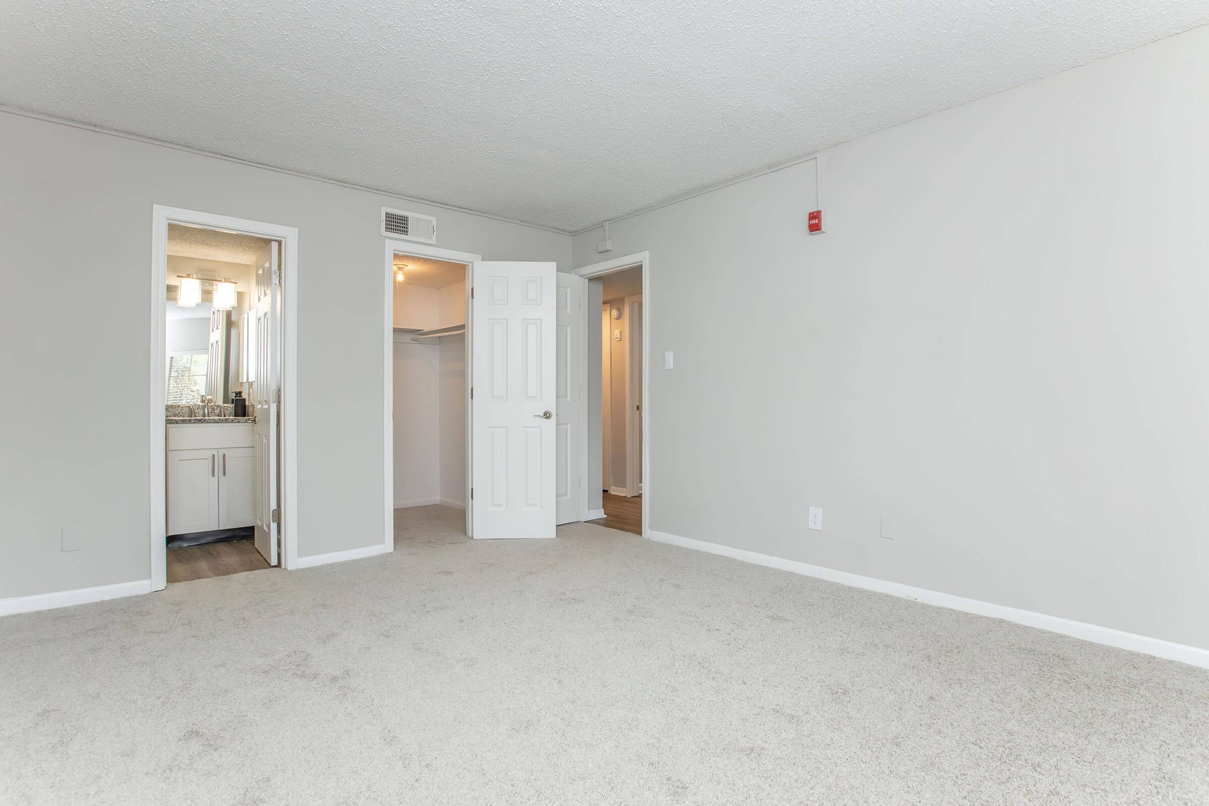 A spacious, empty room with light gray walls and carpeted floor. There are two doorways on the left leading to a bathroom and a closet, with soft lighting visible through the doorways. The room appears bright and inviting, with no furniture present.