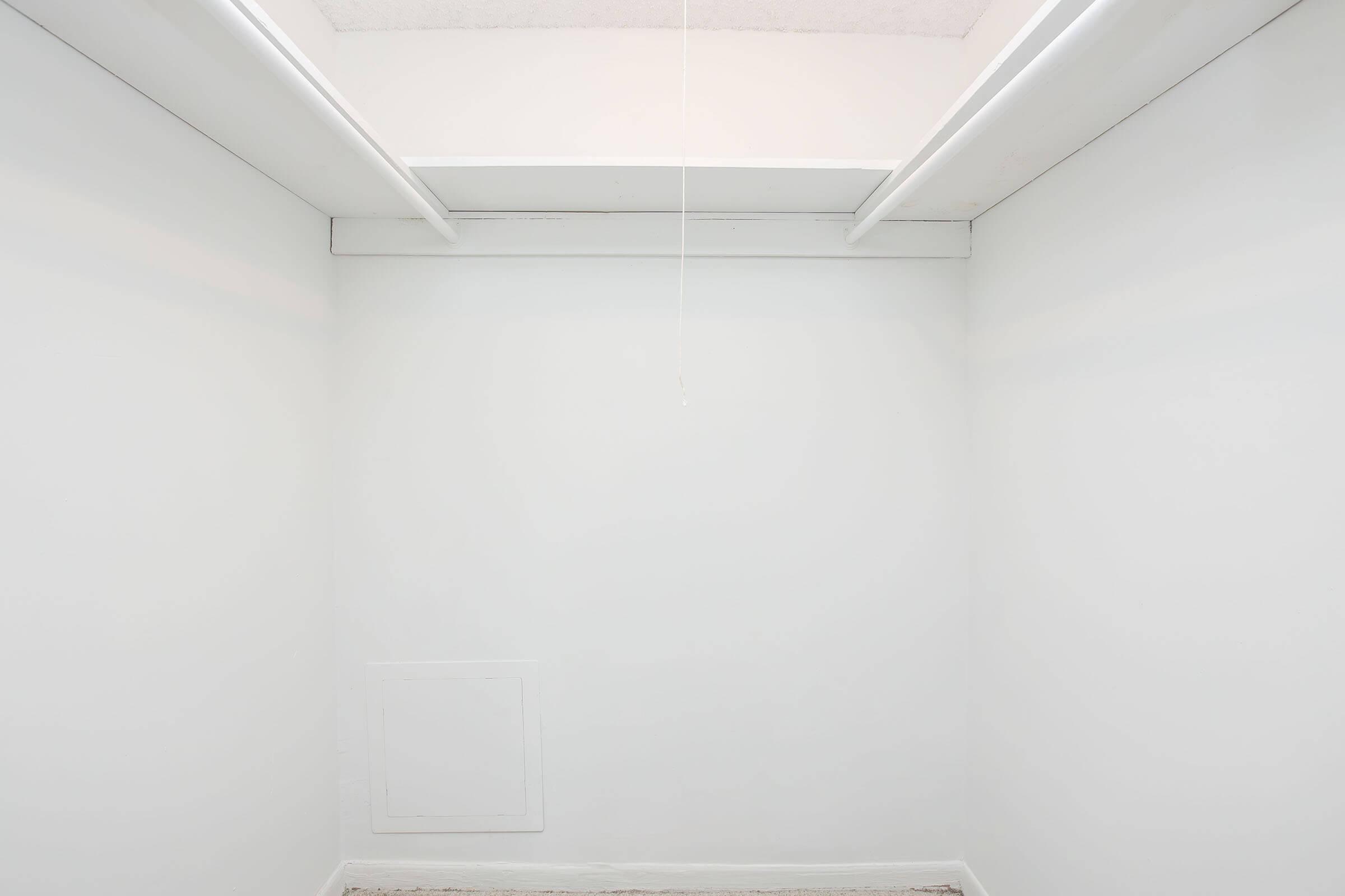 A minimalistic interior of a small, empty room with white walls and a ceiling featuring a vent. A pull cord hangs from the ceiling, and there is a small wall panel or door in the lower left corner. The floor is bare and unobstructed, contributing to the clean and simple aesthetic.