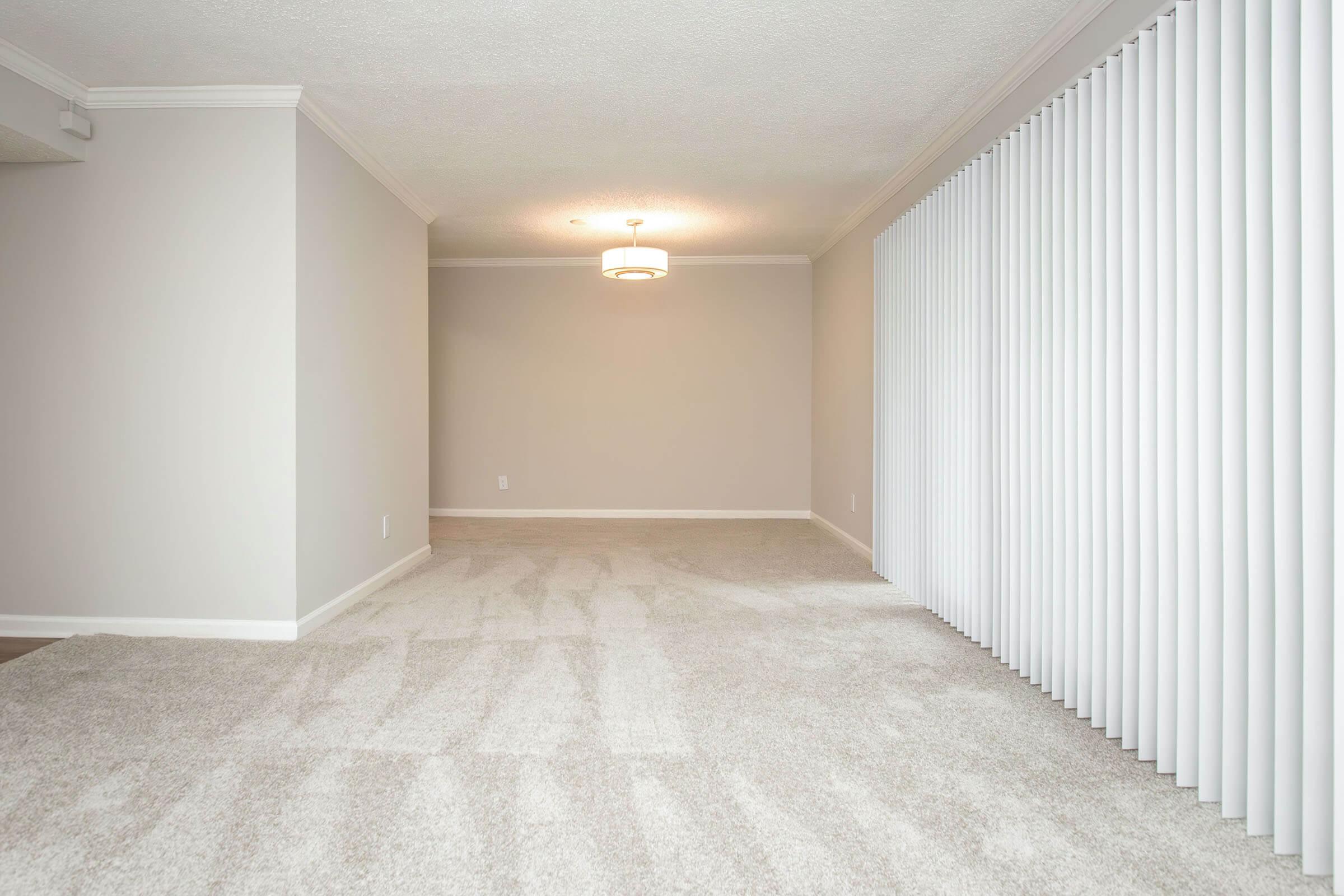 A spacious, empty interior room featuring light gray walls and a plush carpet. The room includes a modern light fixture hanging from the ceiling and a wall of white vertical blinds, providing a bright, inviting atmosphere. Ideal for various living arrangements or decor.