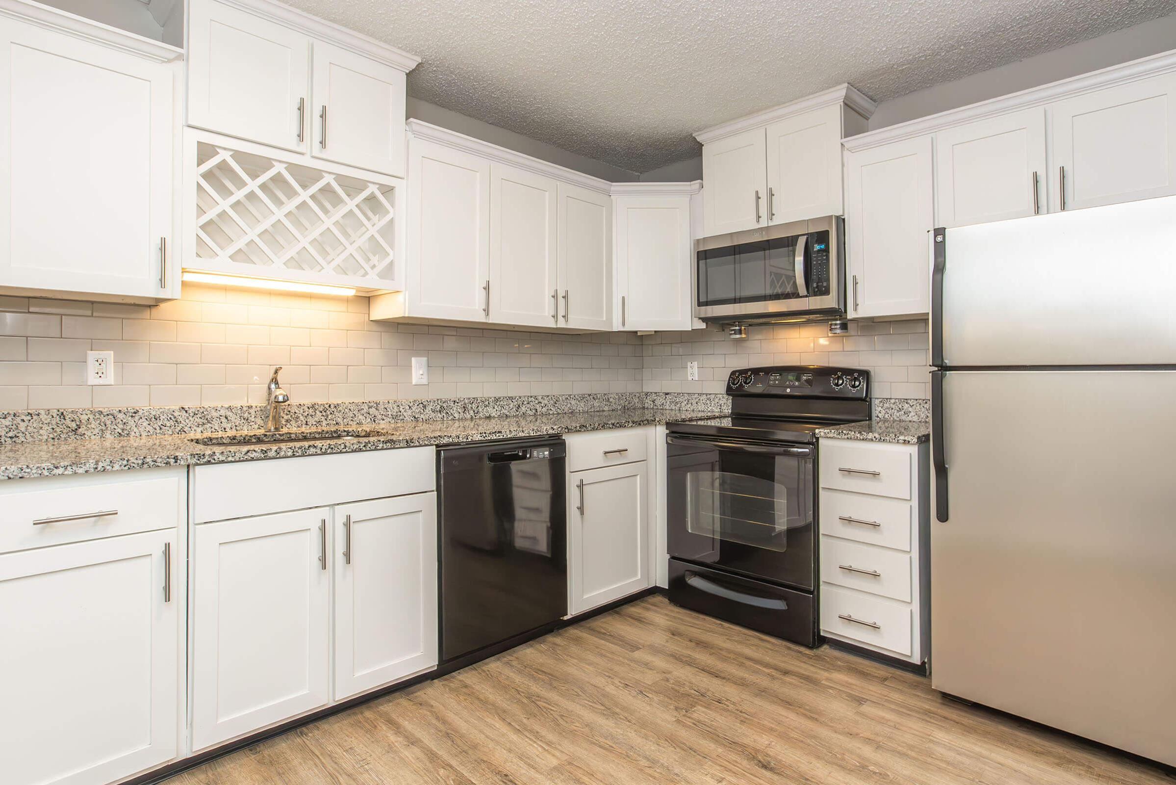 Modern kitchen featuring white cabinets, a granite countertop, stainless steel appliances including a microwave, oven, and refrigerator, and a dishwasher. The flooring is a warm wood tone, creating a cozy yet contemporary atmosphere. The layout is functional and well-organized for cooking and meal preparation.