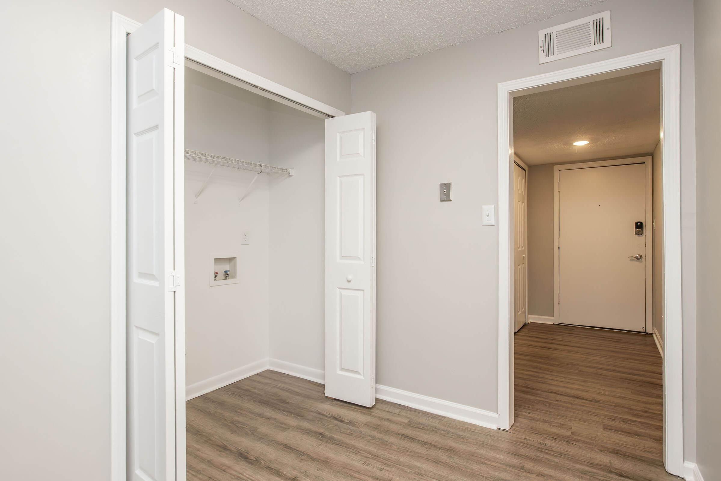 A view of an interior hallway featuring a closet with sliding doors on the left, an empty wall with a built-in shelf, and another door leading to the outside or another room on the right. The walls are painted light gray, and the floor is light wood laminate.