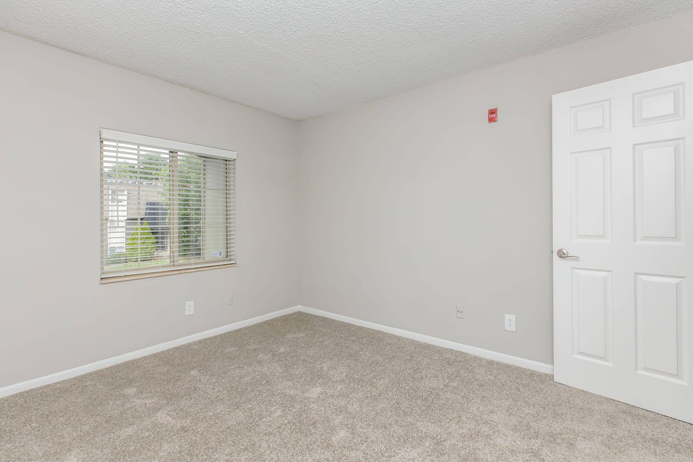 A vacant room with light beige walls and a neutral carpeted floor. There's a small window with blinds allowing natural light, and a closed white door on the right side. The room has a clean, minimalist appearance with no furniture or decorations present.